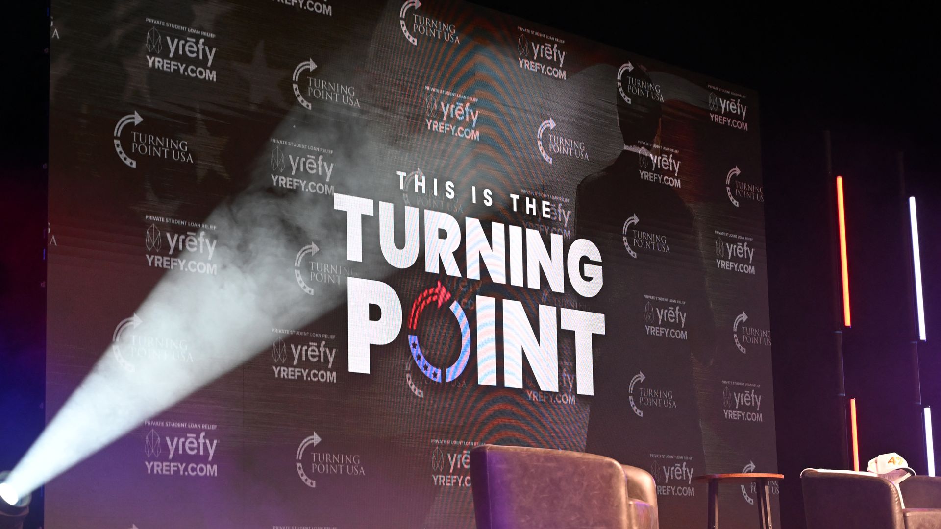A backdrop reads "this is the turning point" with two empty chairs on a stage 