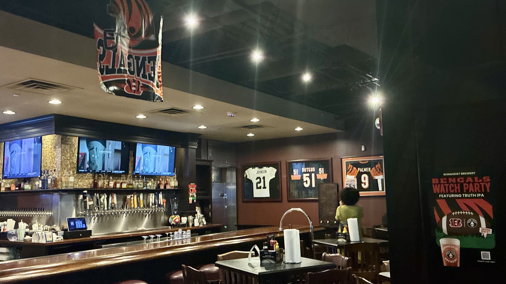 Dimly lit sports bar with a long wooden bar and stools. Multiple TVs line the wall; framed football jerseys (numbers 21, 51, 9) hang above. A red banner dangles from the ceiling.