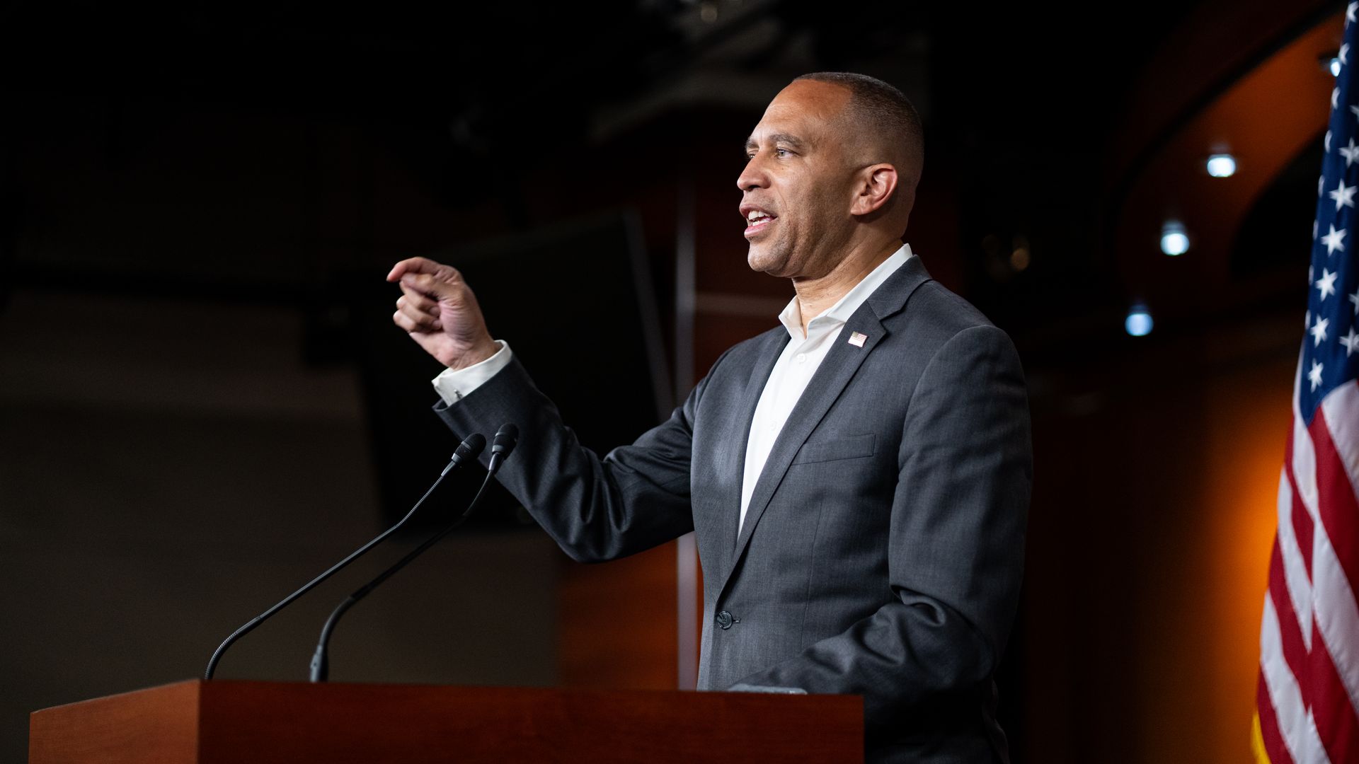 Hakeem Jeffries, wearing a gray suit and speaking at a podium in front of an American flag.