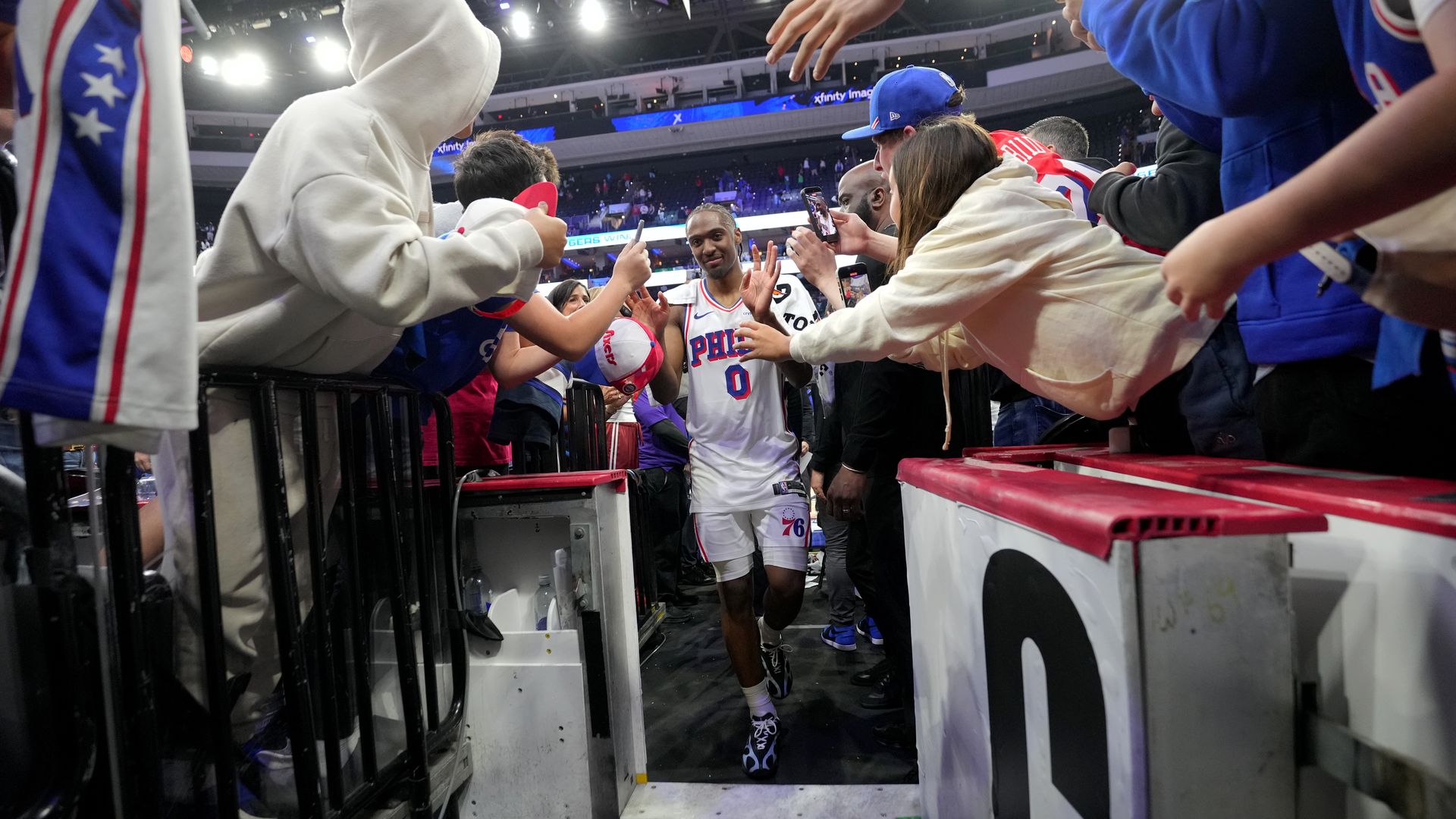 Basketball player in a white Philadelphia 76ers jersey with number 0 walks through a tunnel as excited fans lean in, phones out, crowd noise around, bright arena lights overhead.