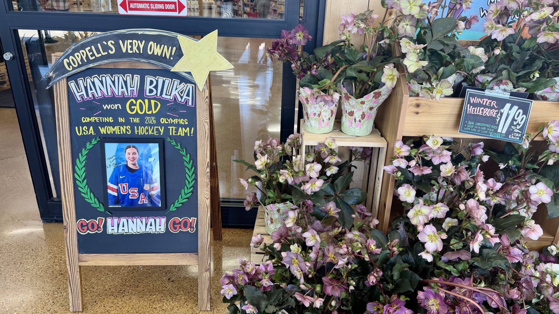 Wooden chalkboard sign in front of store celebrating Hannah Bilka winning gold for USA women's hockey team 2026 Olympics, next to shelves of pink and white winter hellebore flowers.