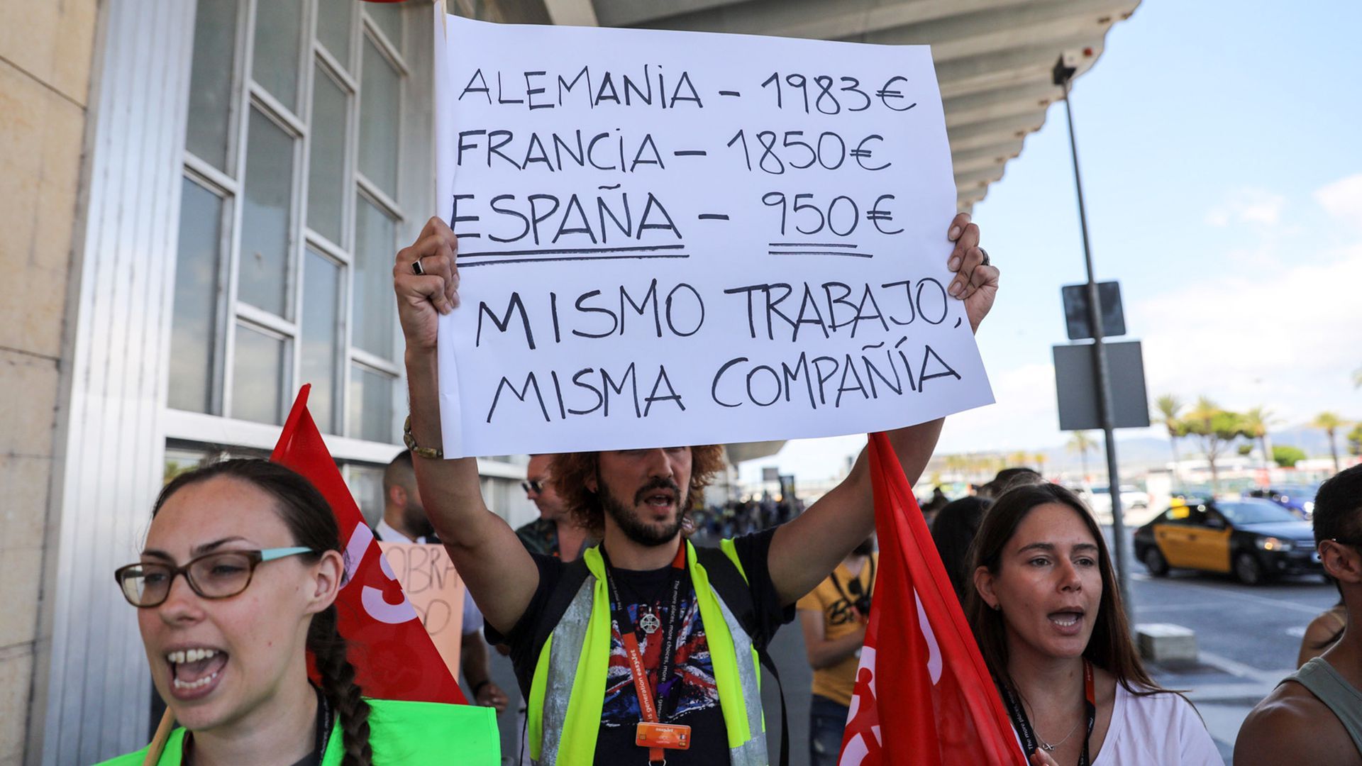 striking workers hold up a sign that in Spanish says how much they're getting paid in different countries