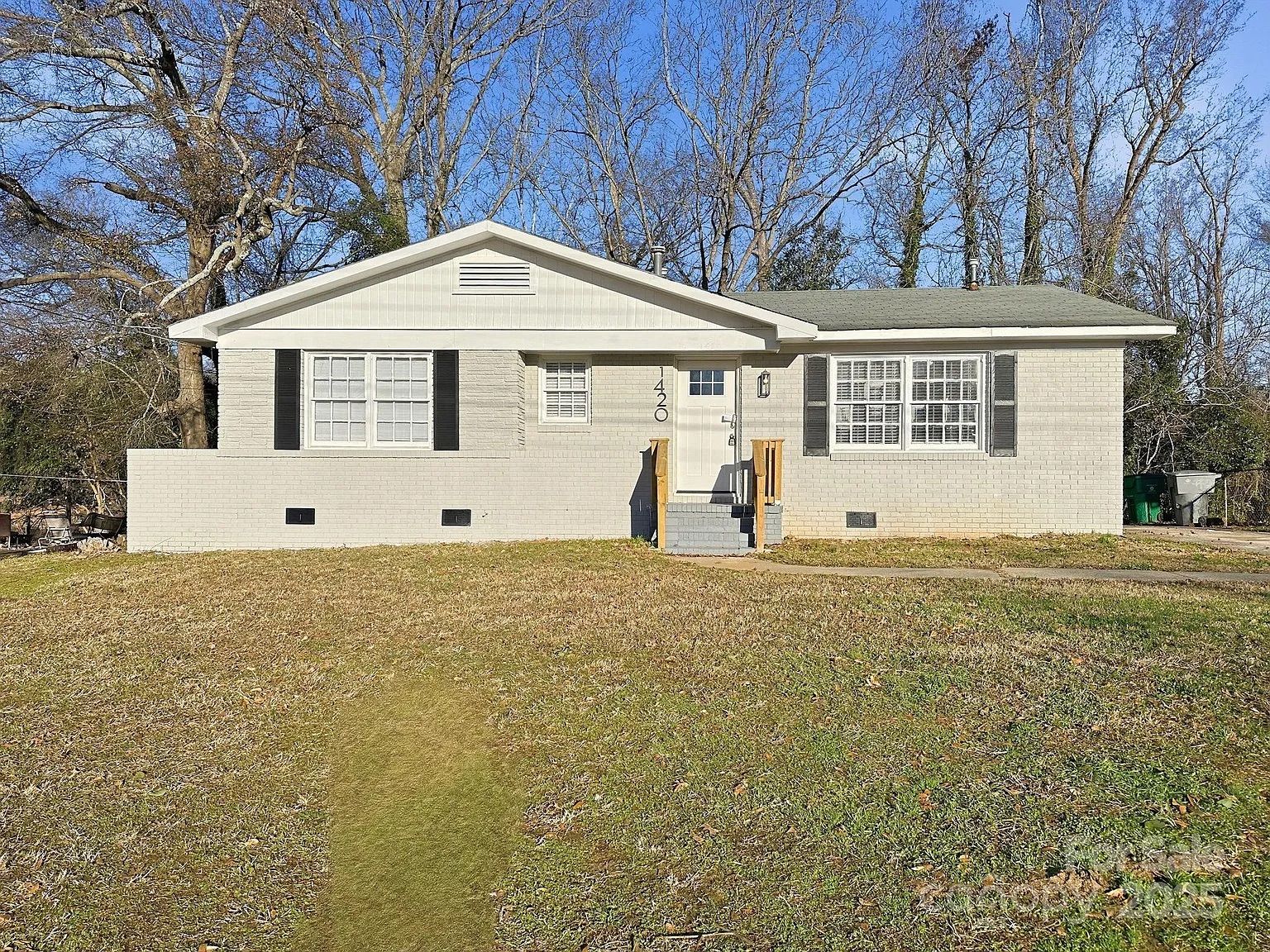 Single-story beige brick house with white front door, black shutters, and gray roof, set against bare trees and a blue sky, with a grass lawn in front.