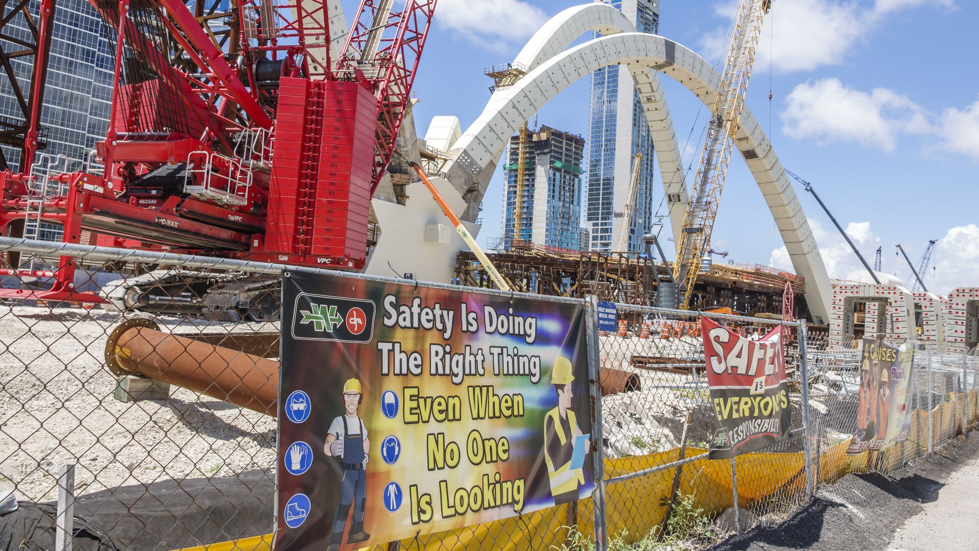 Chain-link construction fencing with work-safety banners around construction equipment next to the I-395 Signature Bridge project in downtown Miami.