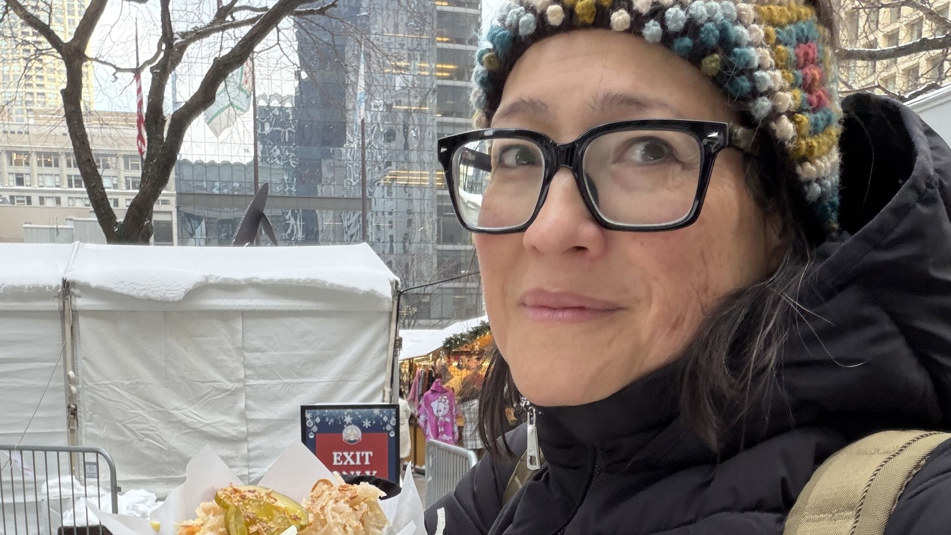 Woman wearing glasses, a colorful knit hat, and a black jacket holds food with pickles and shredded topping outdoors at a winter market with snow and tents in the background.