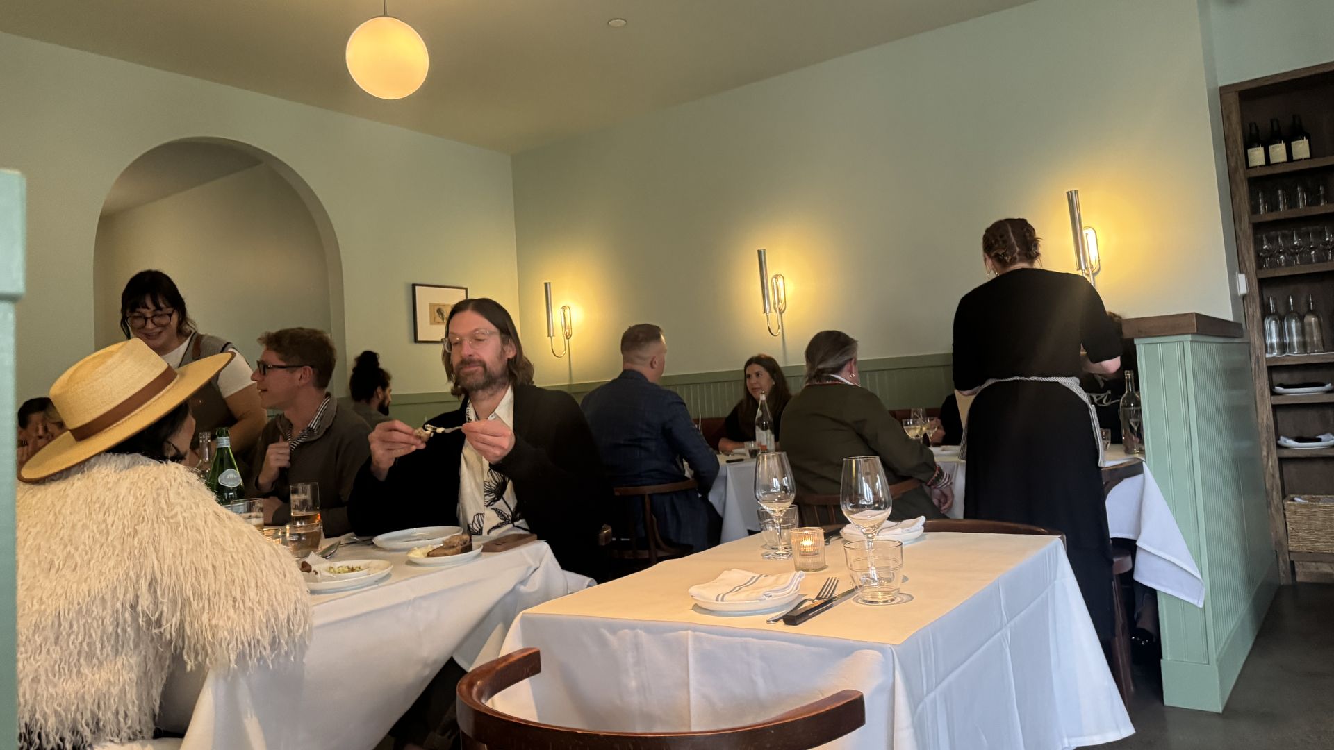 Cozy restaurant scene with diners at tables covered in white cloths. Soft yellow wall lights, a round ceiling light, and wooden chairs create warm ambiance. A woman in a straw hat and fluffy shawl is visible.