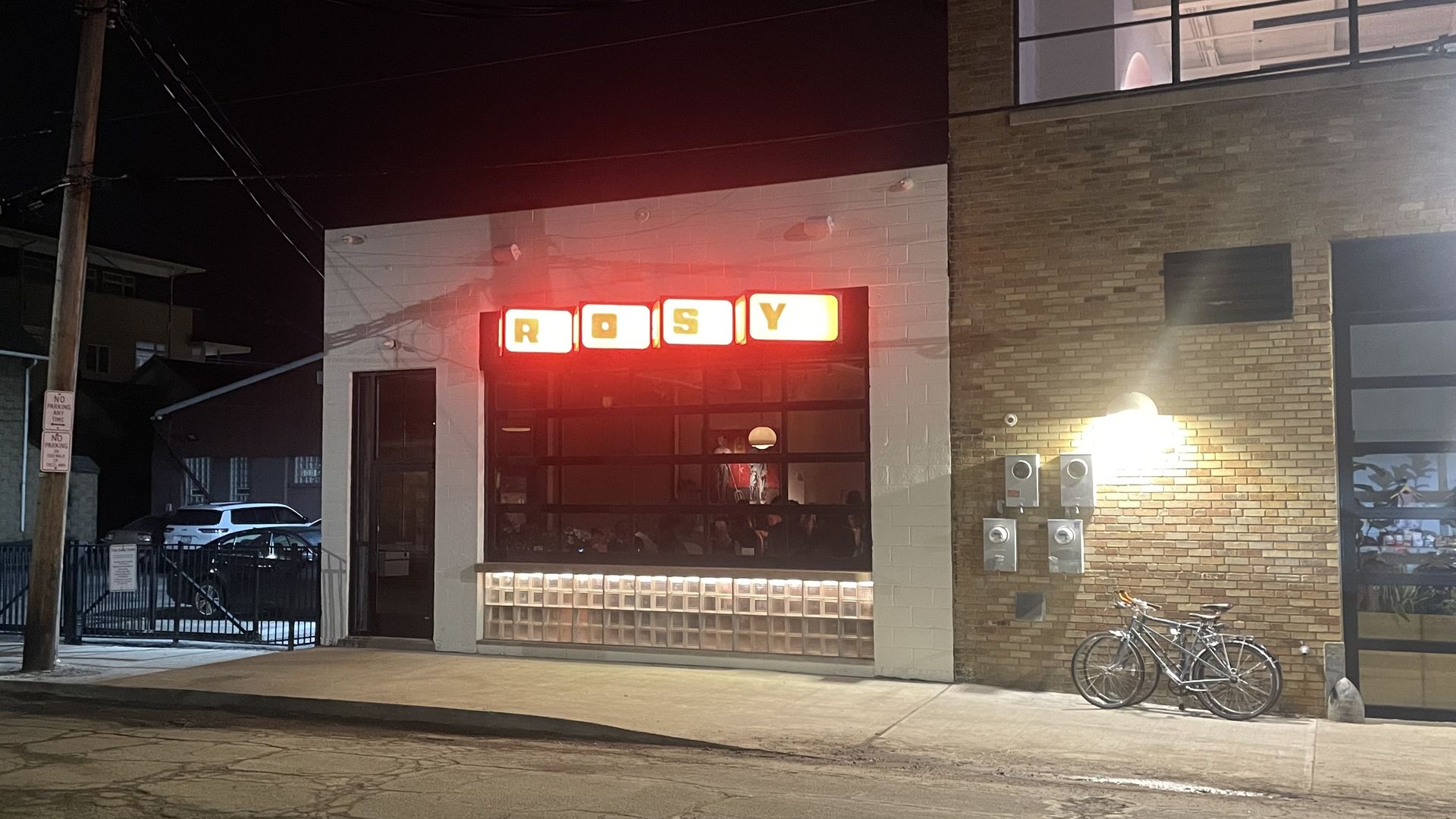Night scene of a brick and white building with red neon sign reading "ROSY" above a large window, a bicycle parked by a lit wall, and parked cars behind a gated area.