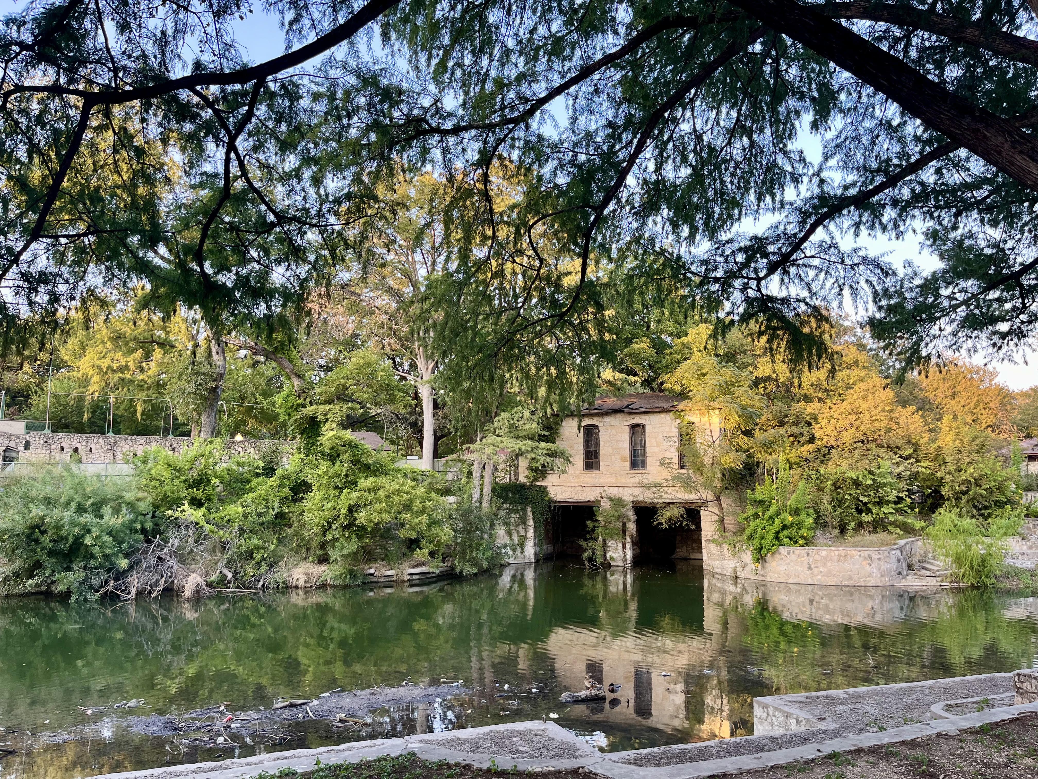 The Pump House in Brackenridge Park is an old building seen across the river and through large trees.