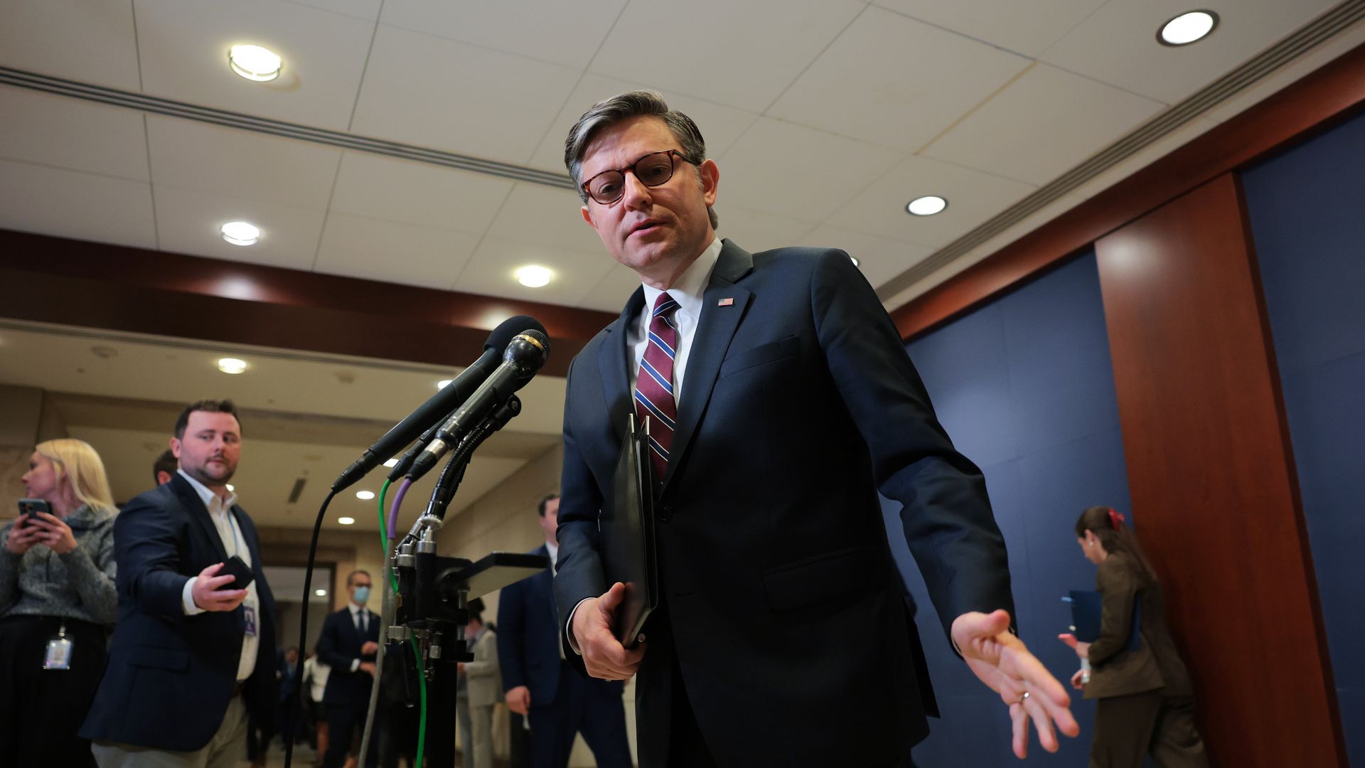 Man in a dark suit, striped tie, and glasses standing behind a microphone stand speaking while holding a folder, with people in business attire in the background in a conference hallway.