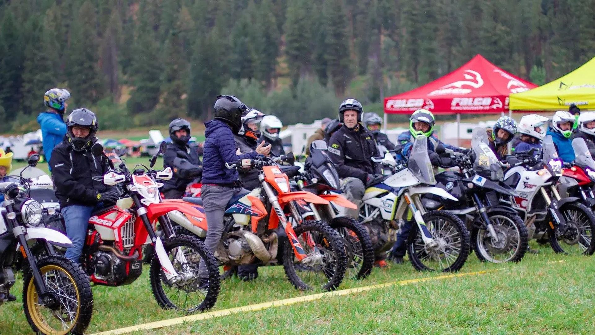 A line of motorcycle riders in helmets on a grassy field, with tents behind.