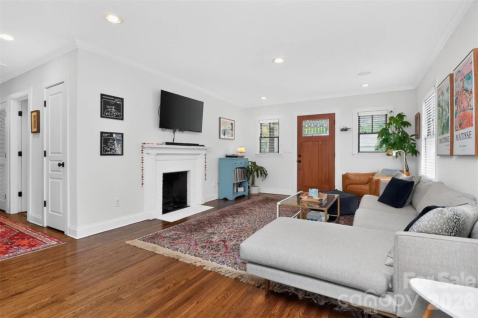 Bright living room with white walls and hardwood floors, a wall-mounted TV above a white fireplace, blue cabinet, large plant, gray sectional sofa, and framed Matisse posters by the window and door.