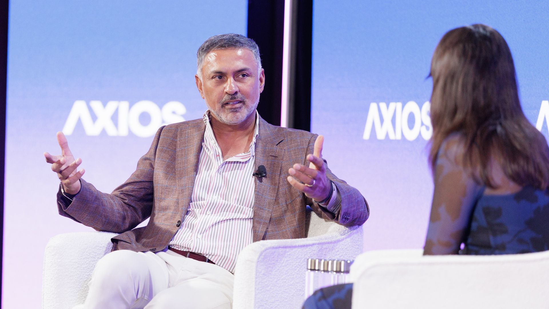 Two people in conversation during an Axios event, man in brown blazer and white pants gesturing as he speaks, woman with long hair listens, blue backdrop with Axios logo.