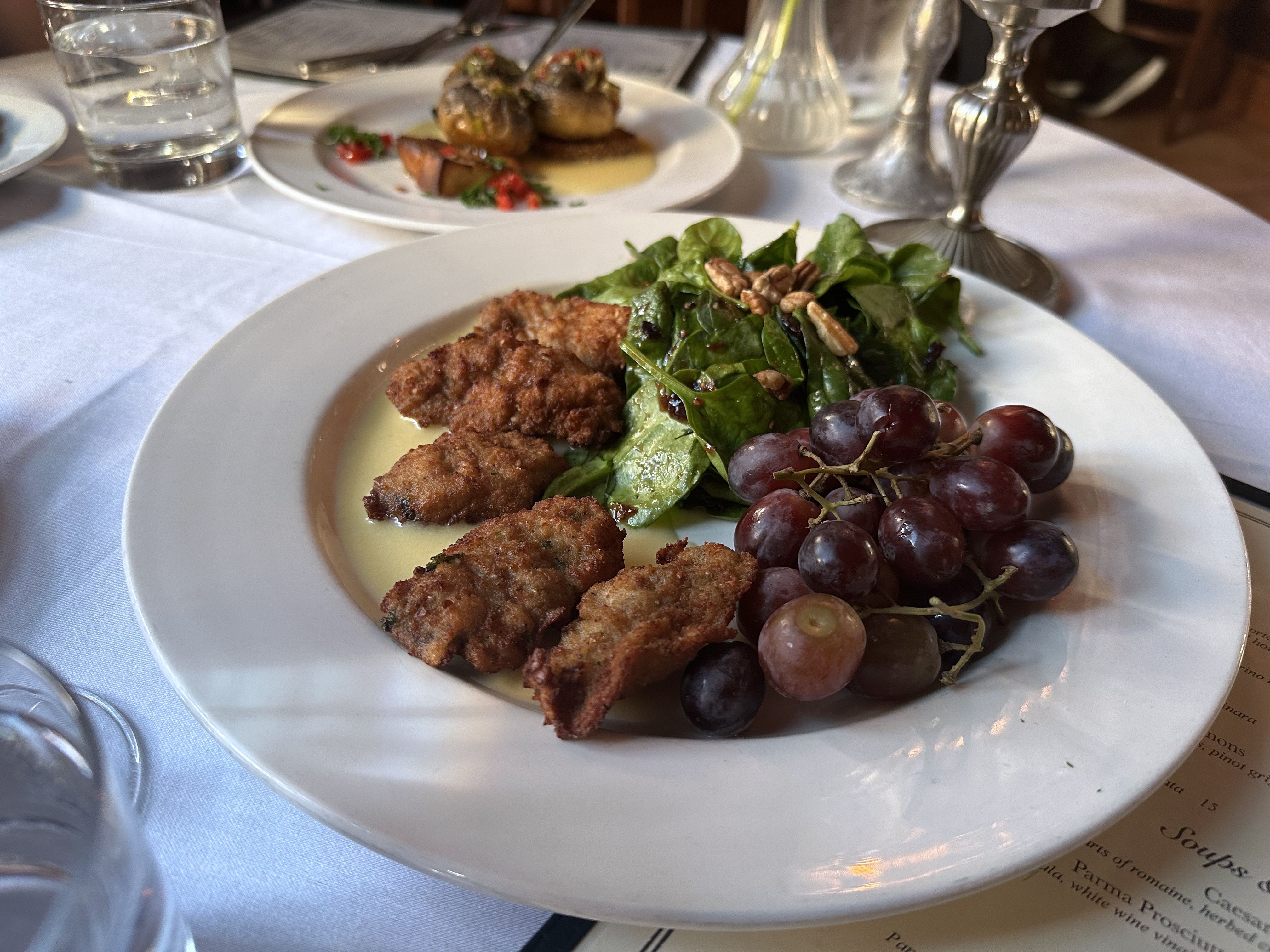 Four fried oysters sit on a large white plate atop a cream sauce. Next to the oysters is a pecan-topped spinach salad and red grapes.