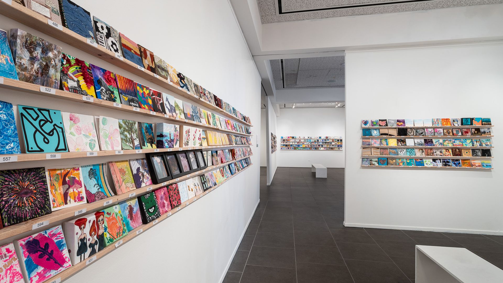 A displays of hundreds of colorful 5x5 art pieces on a wall of an art gallery.