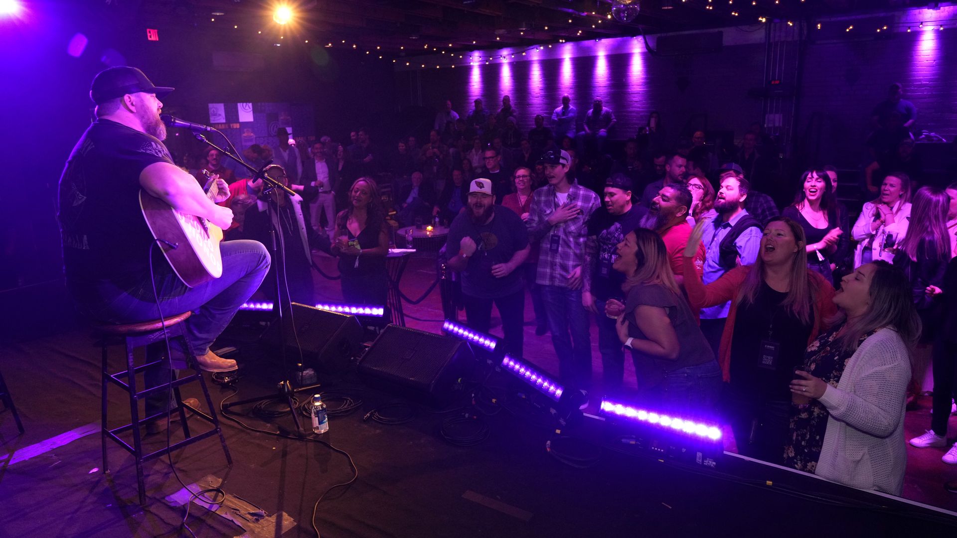 Bearded man wearing a cap plays acoustic guitar and sings on stage with purple lighting as a lively audience watches and cheers in a dimly lit venue with string lights on the ceiling.