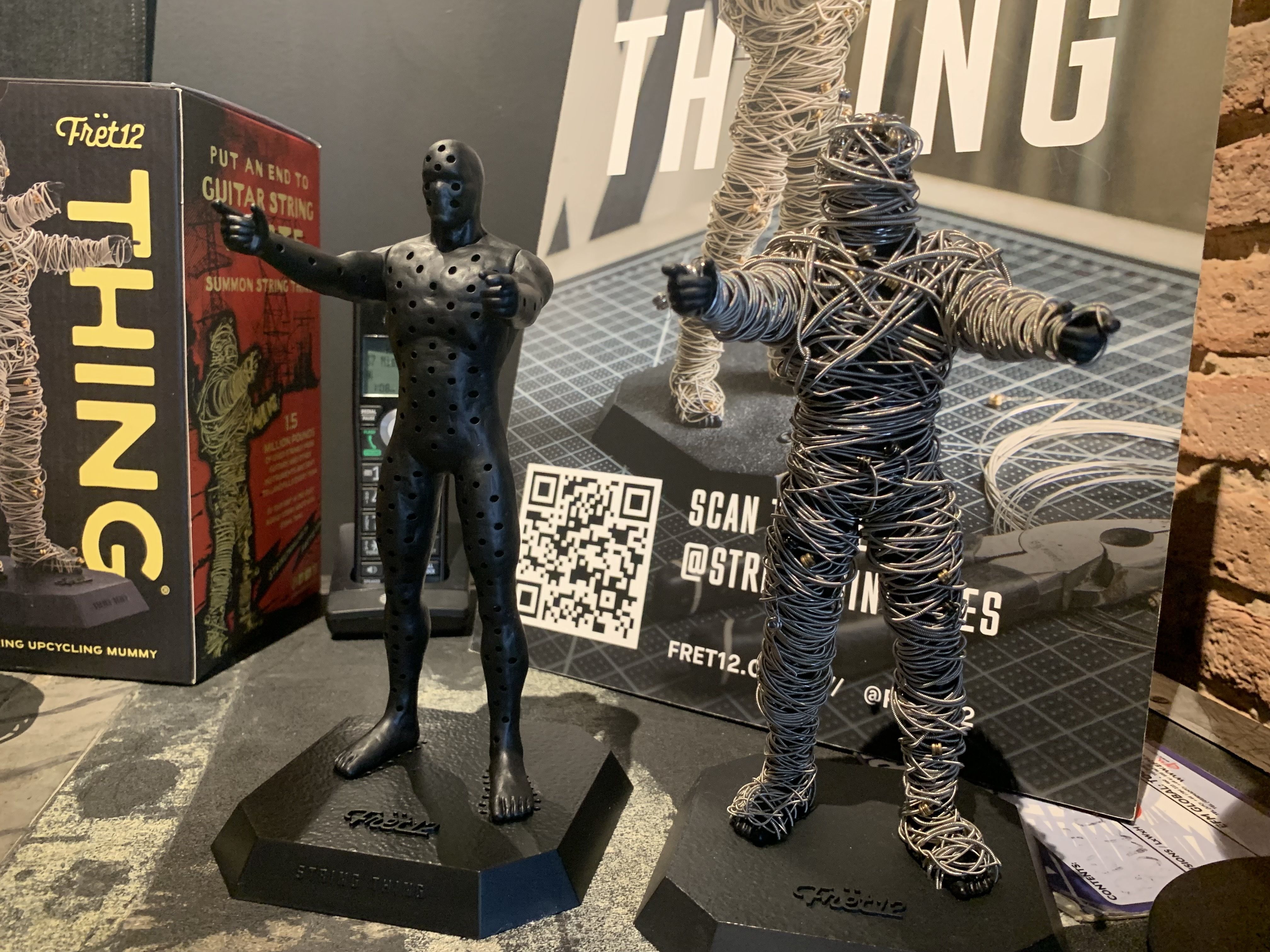 Two black statuettes of human figures on octagonal bases; left one solid with holes, right one wrapped in silver wire, displayed with boxes and promotional materials in background.