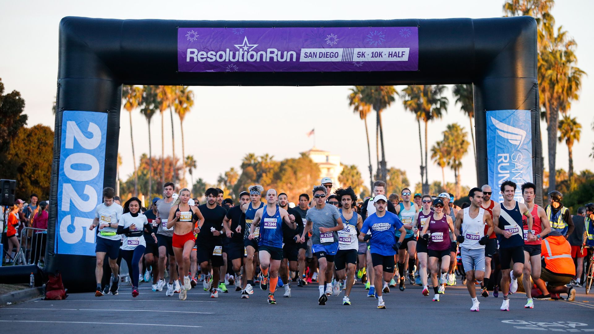 Runners start Resolution Run 2025 in San Diego under a black inflatable arch with purple sign, palm trees and buildings in background, participants in varied athletic attire.