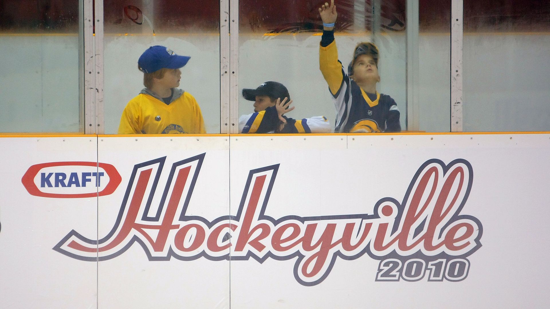 Three children at hockey rink watching through the glass, with a Kraft sign etched into the wall just below them