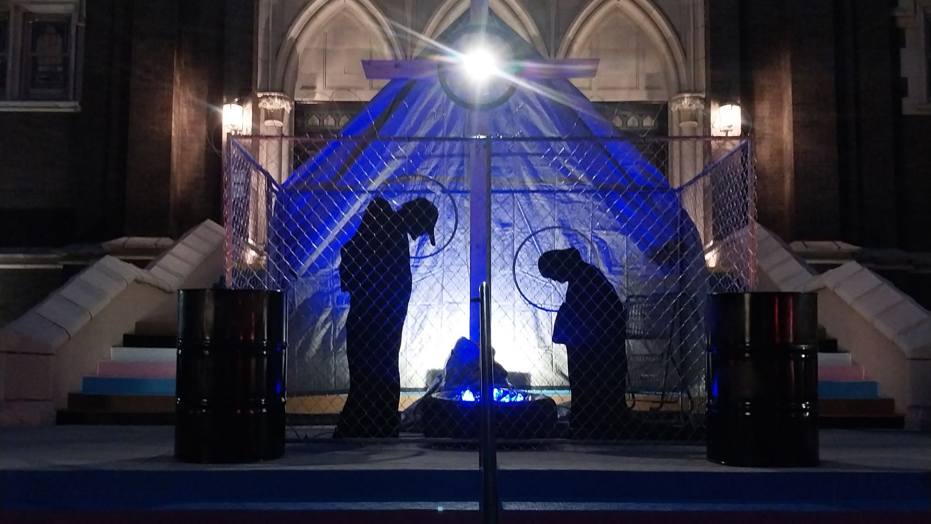 Night scene of two silhouette figures behind chain-link fence in front of blue-lit tarp, with a cross above and bright light shining, set against a building with steps and arched windows.