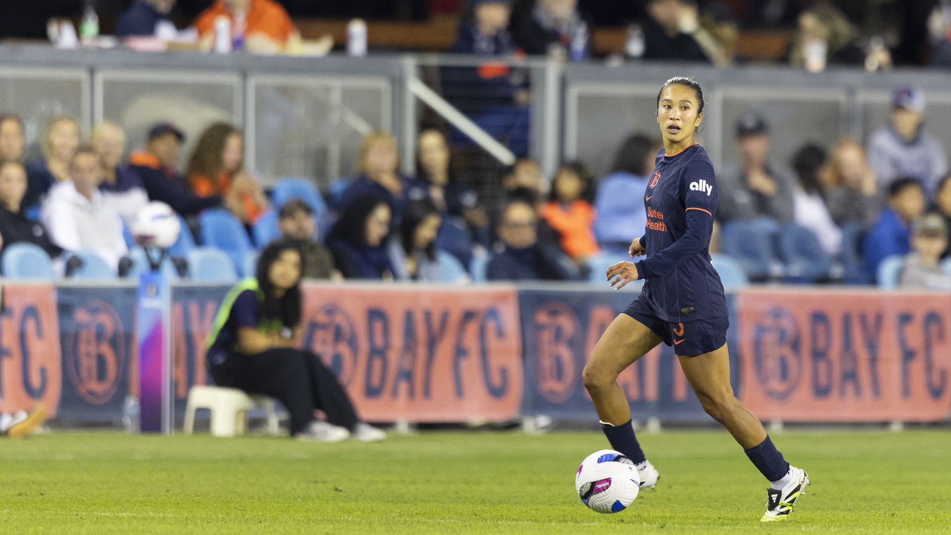Female soccer player in navy uniform with orange text controls soccer ball on green field, with blurred crowd and "BAYFC" banner in background at night game.