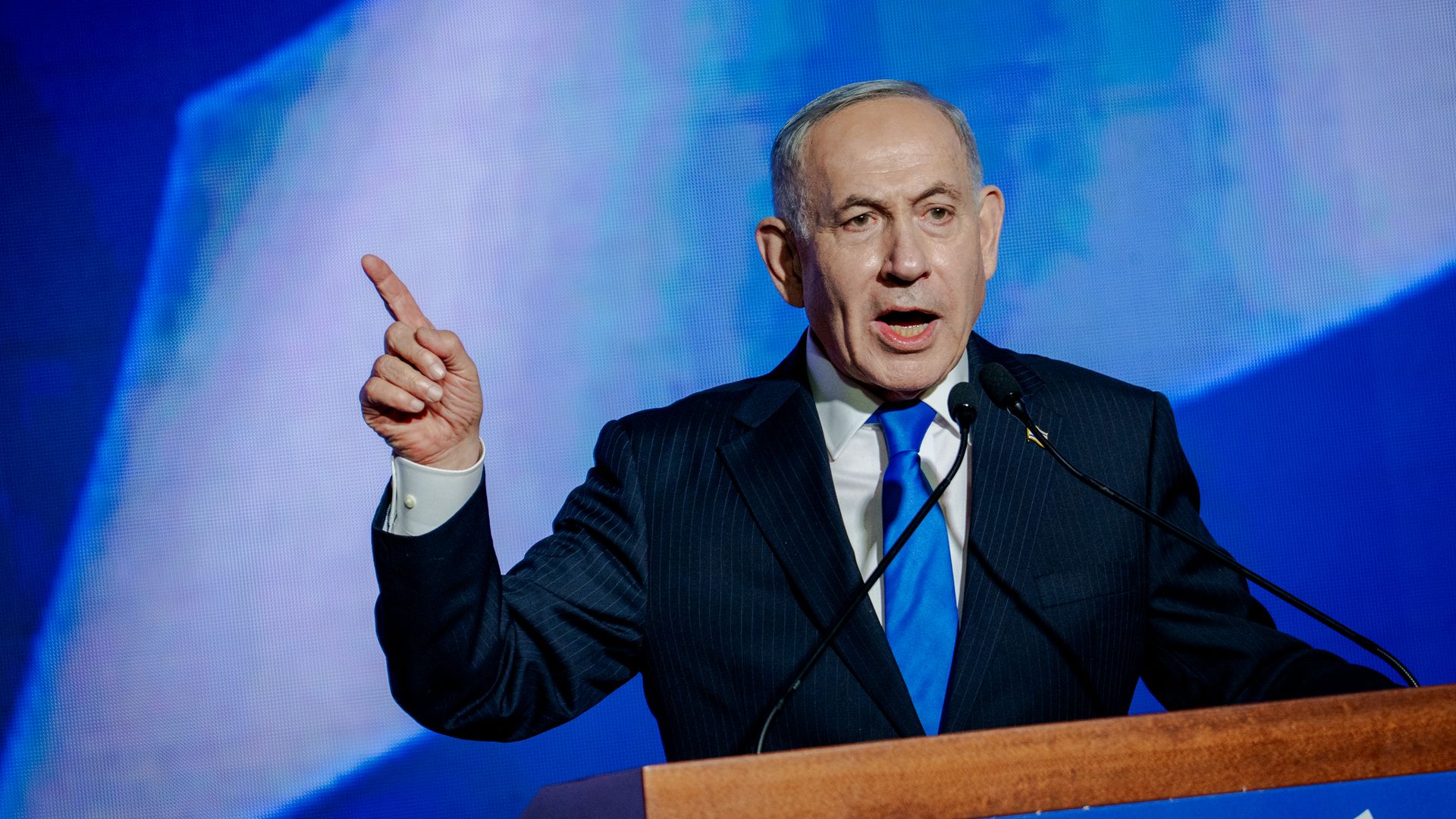 Israeli Prime Minister Benjamin Netanyahu, an older man in a dark pinstripe suit and blue tie, delivers a speech at a wooden podium with microphones, raising one finger as he speaks, against a blue abstract background.