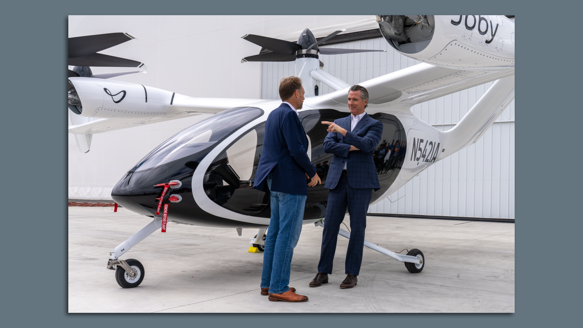 Image of California Gov. Gavin Newsom chatting with Joby Aviation CEO JoeBen Bevirt next to Joby's first electric vertical takeoff and landing aircraft, which looks like a helicopter, but with multiple small rotors. 