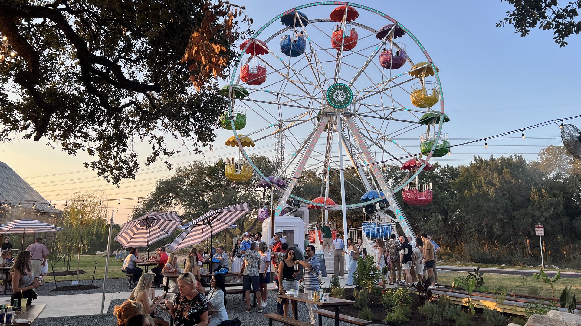 Outdoor scene with a colorful Ferris wheel, people socializing at picnic tables under striped umbrellas during sunset, surrounded by trees and string lights.