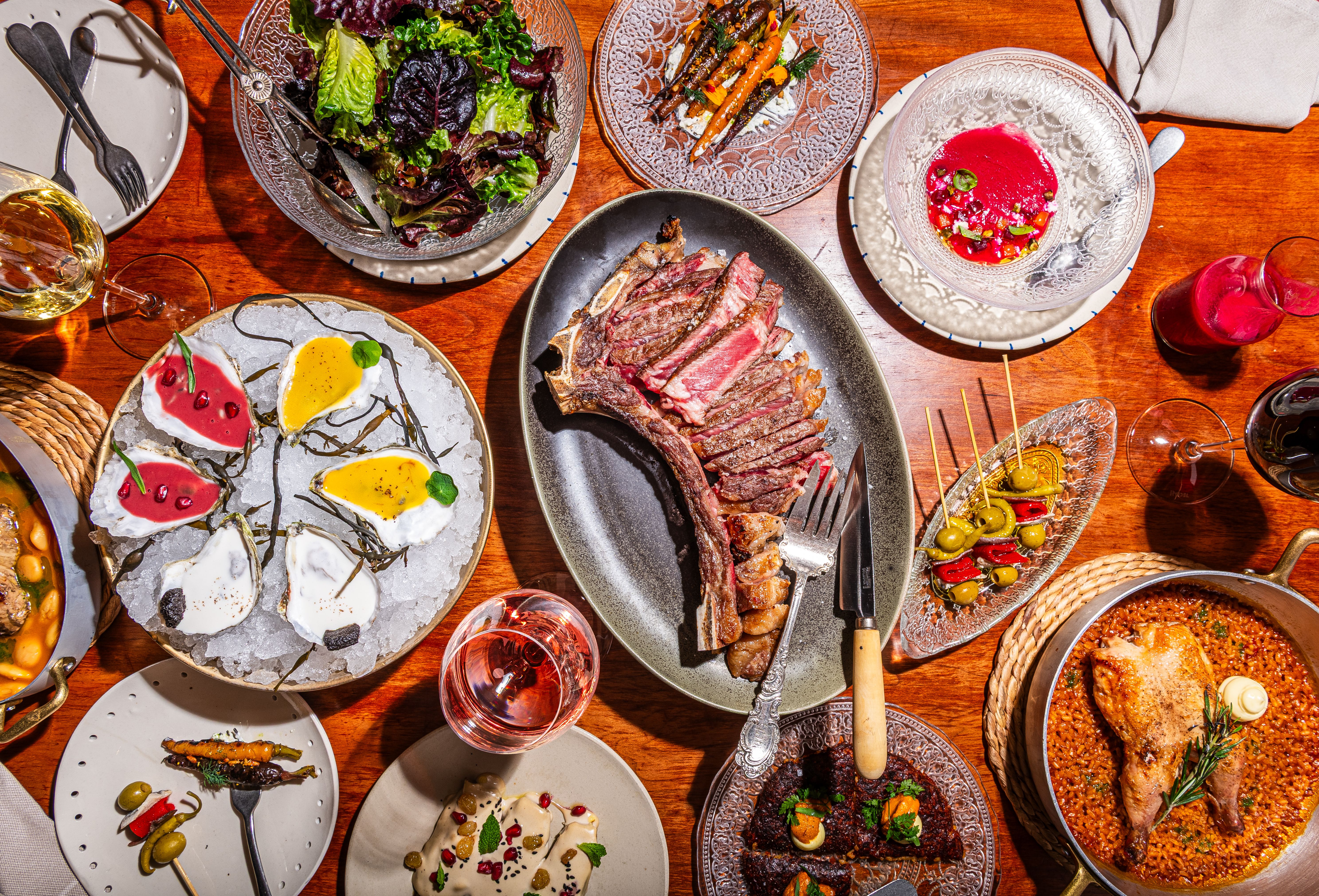 Top-down view of a wooden table laden with a feast: sliced steak on a dark plate, mixed greens salad, oysters on ice with sauces, roasted vegetables, a beet soup, paella, and wine glasses.