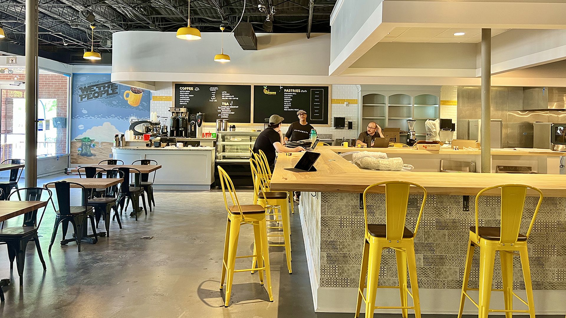 Inside a coffee shop, therer are yellow stools lined up around the counter space and a mural in the back that reads "we the people."