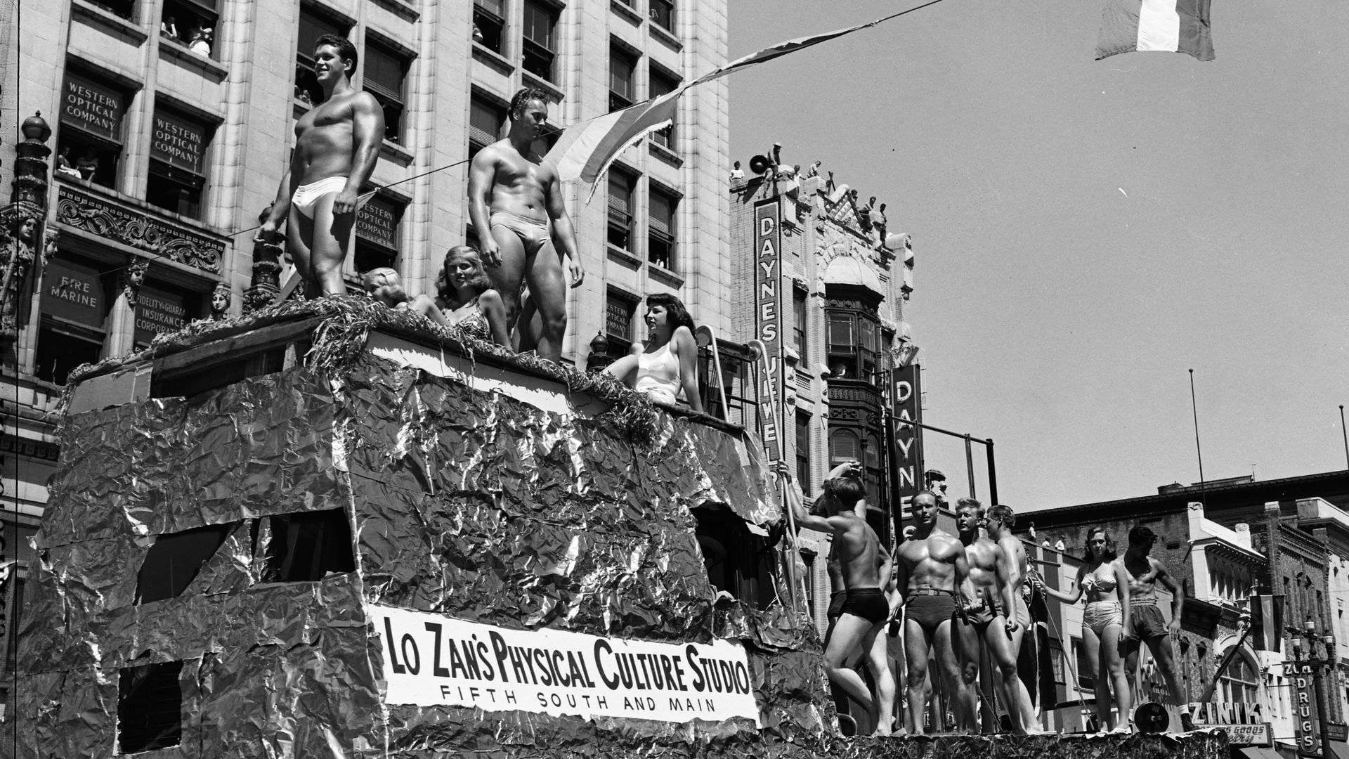 Men in Speedos and body oil stand proudly on a parade float in a black and white photo.
