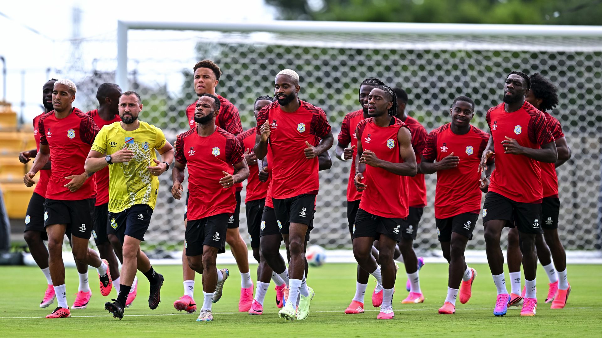 A group of soccer players in red training jerseys run across a green field during a drill, led by a goalkeeper in yellow; a goal net and stands blur in the background.
