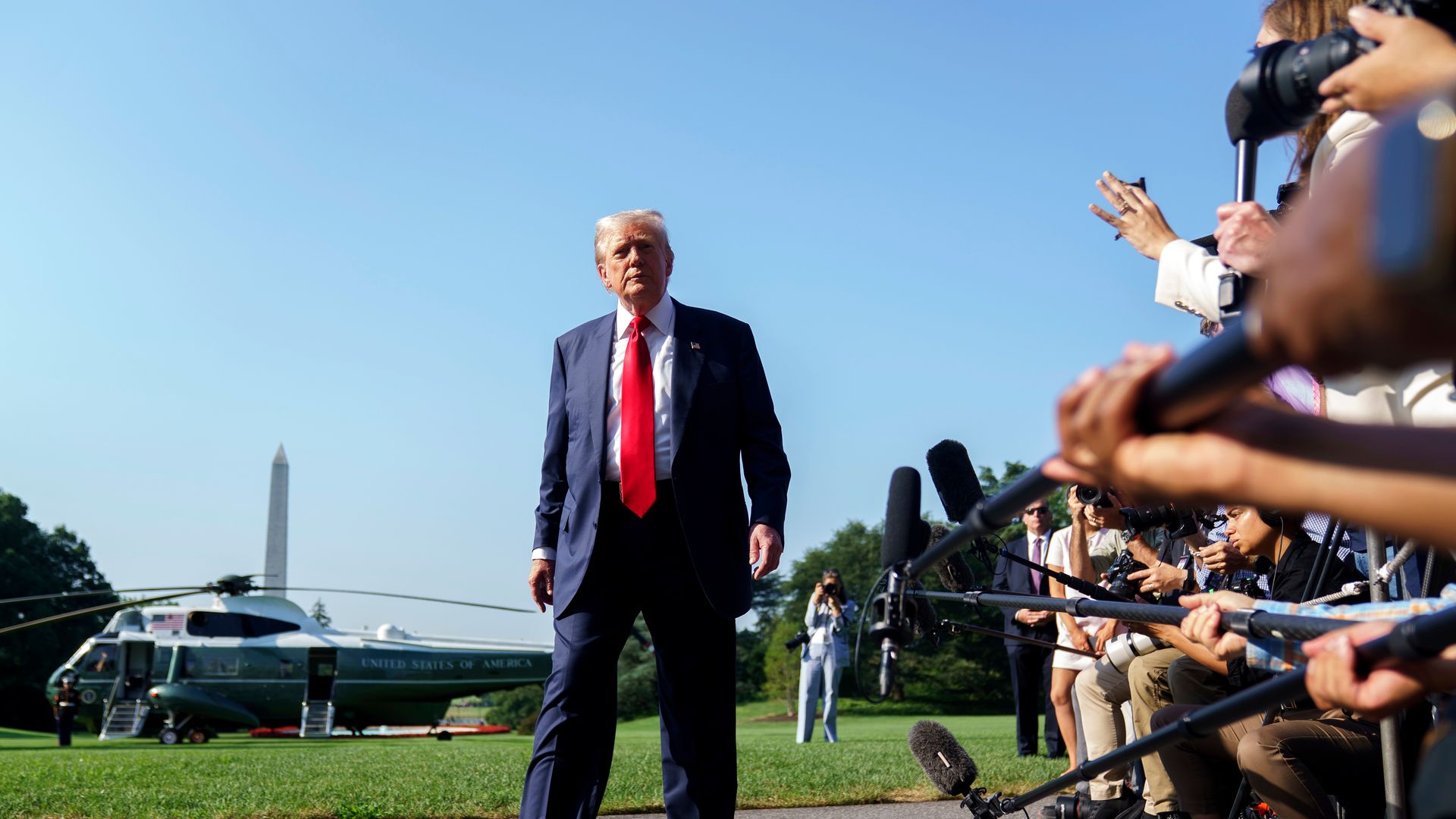 President Trump walking on a path with a helicopter and the Washington Monument in the background, while reporters with microphones and cameras record him.