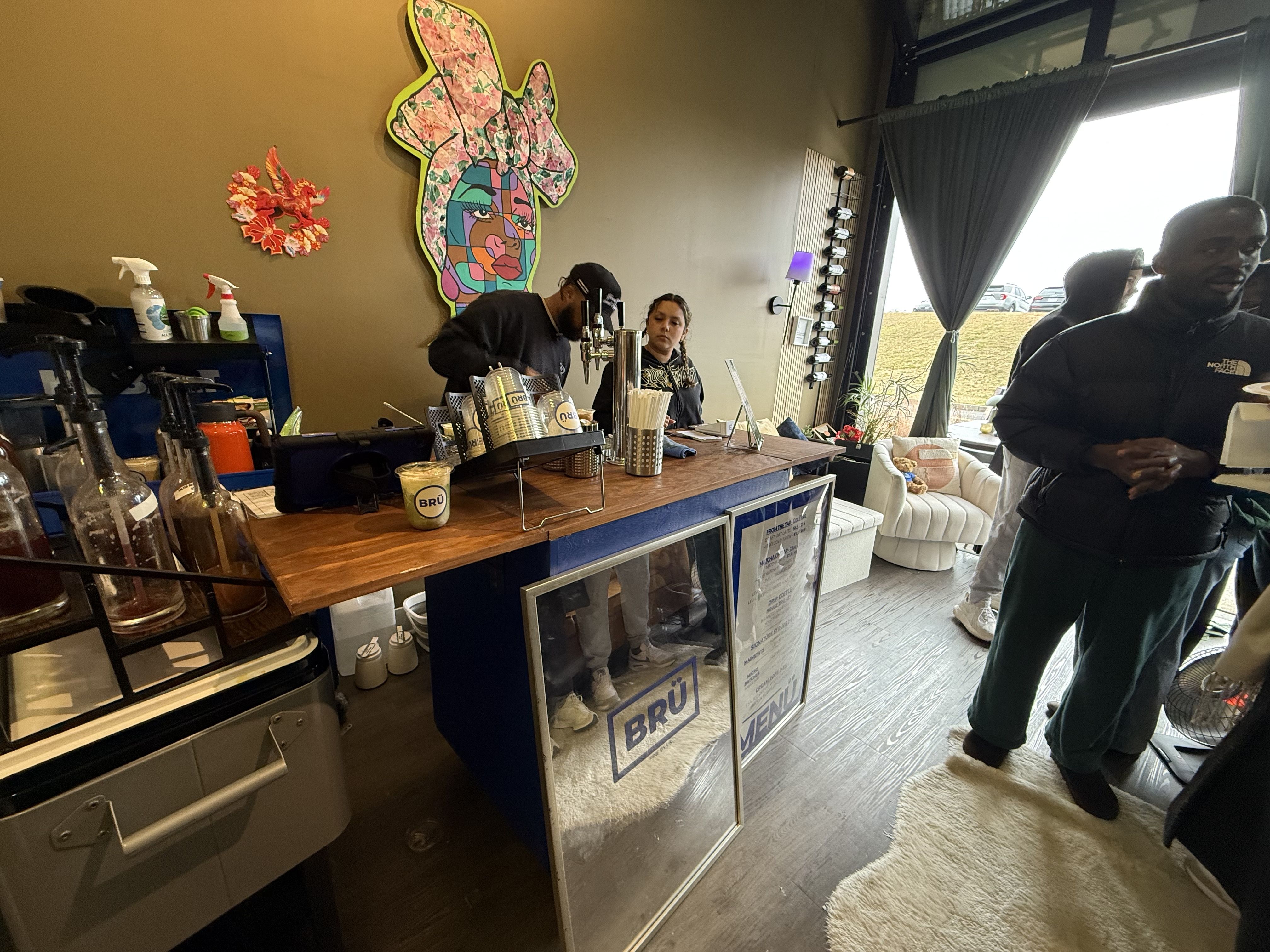Coffee shop interior with people at the counter; colorful abstract woman artwork on the wall; wooden counter with dispensers and cups; window with gray curtains letting in daylight.