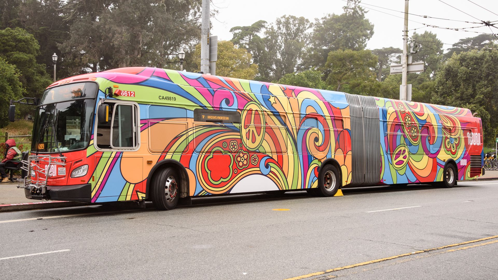 A colorful articulated bus covered in a vibrant psychedelic pattern with swirls, flowers, and peace signs, parked on a street with trees in the background.