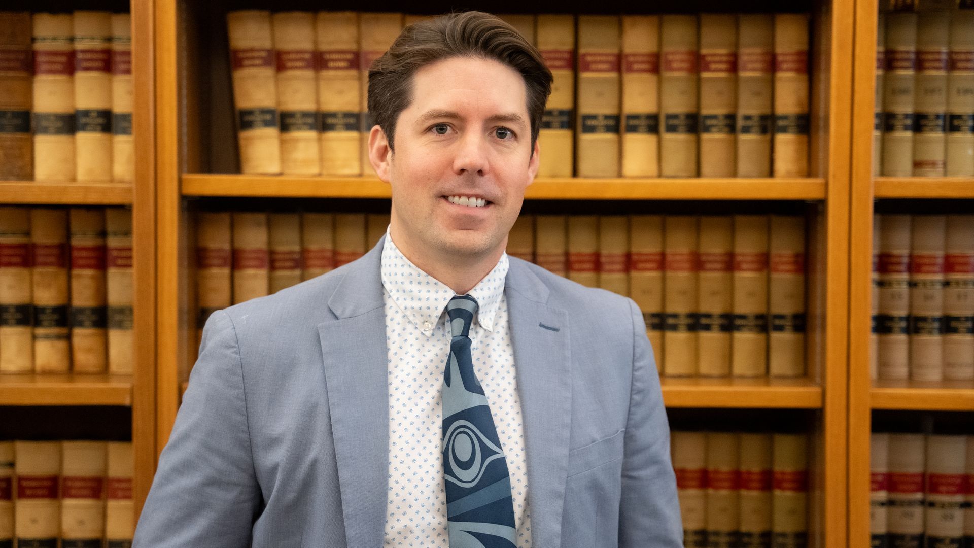 Mike Webb is shown wearing a light blue suit jacket, white shirt with blue patterns, and a blue patterned tie, standing in front of shelves filled with brown legal books.
