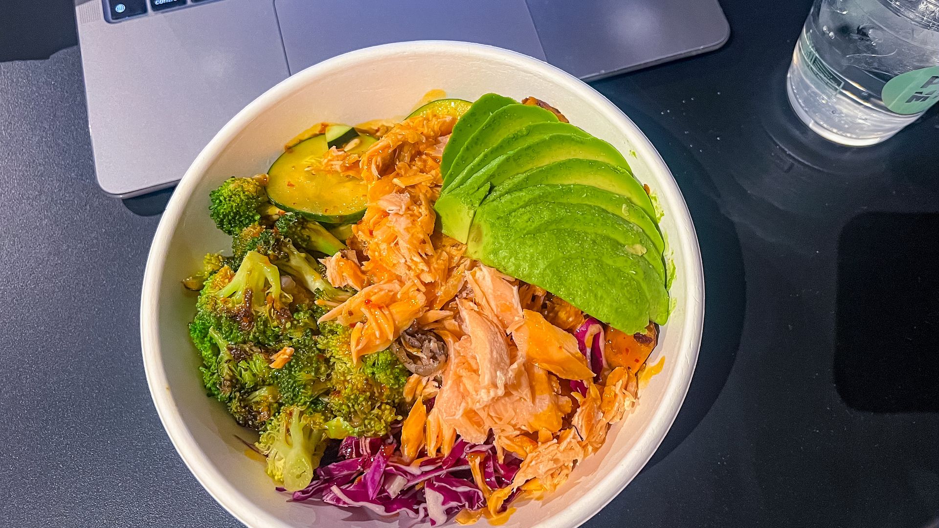 A bowl filled with salmon, avocado, cabbage, broccoli and more sits on a black table with a laptop behind it.