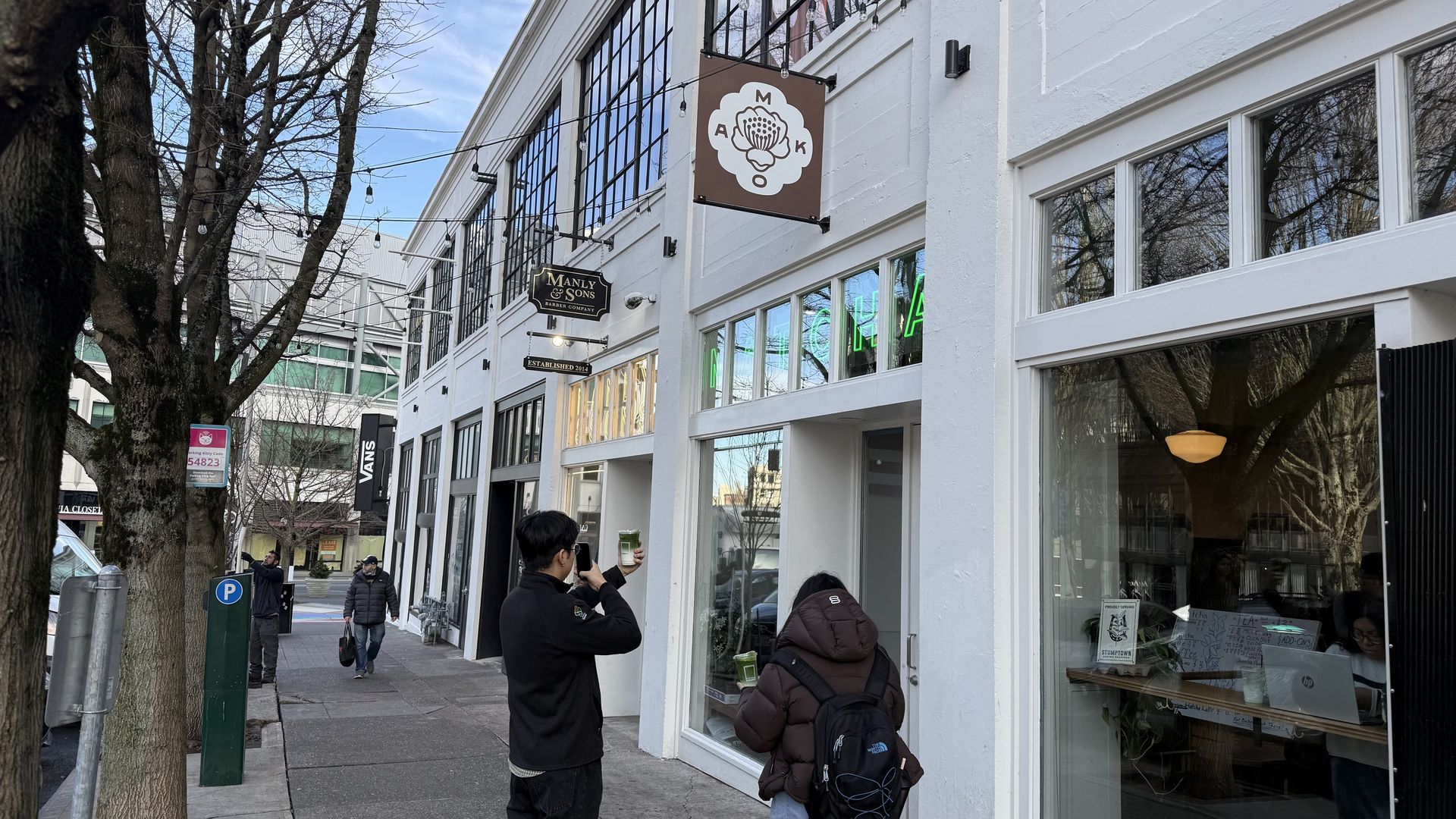 Two people on a city sidewalk near white building with signs for "Manly & Sons" and another with floral logo. One person takes photo of drinks held by the other.