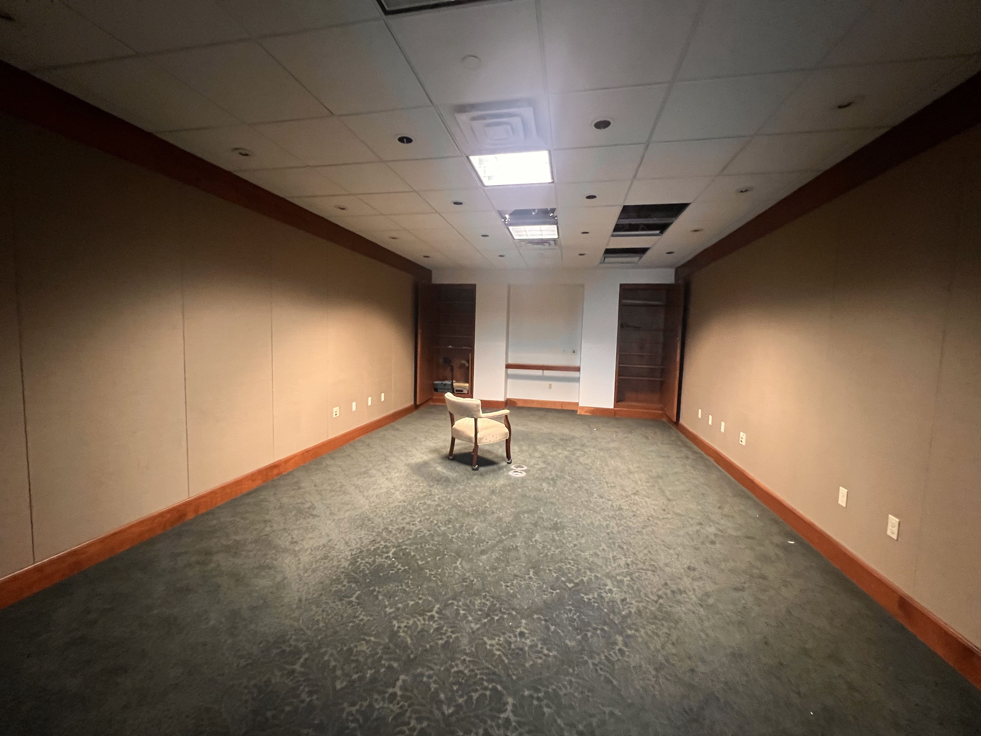 Empty rectangular office space with brown walls, blue-gray carpet, and a white drop ceiling showing missing tiles. A single chair sits in the center; cabinets line the far wall.