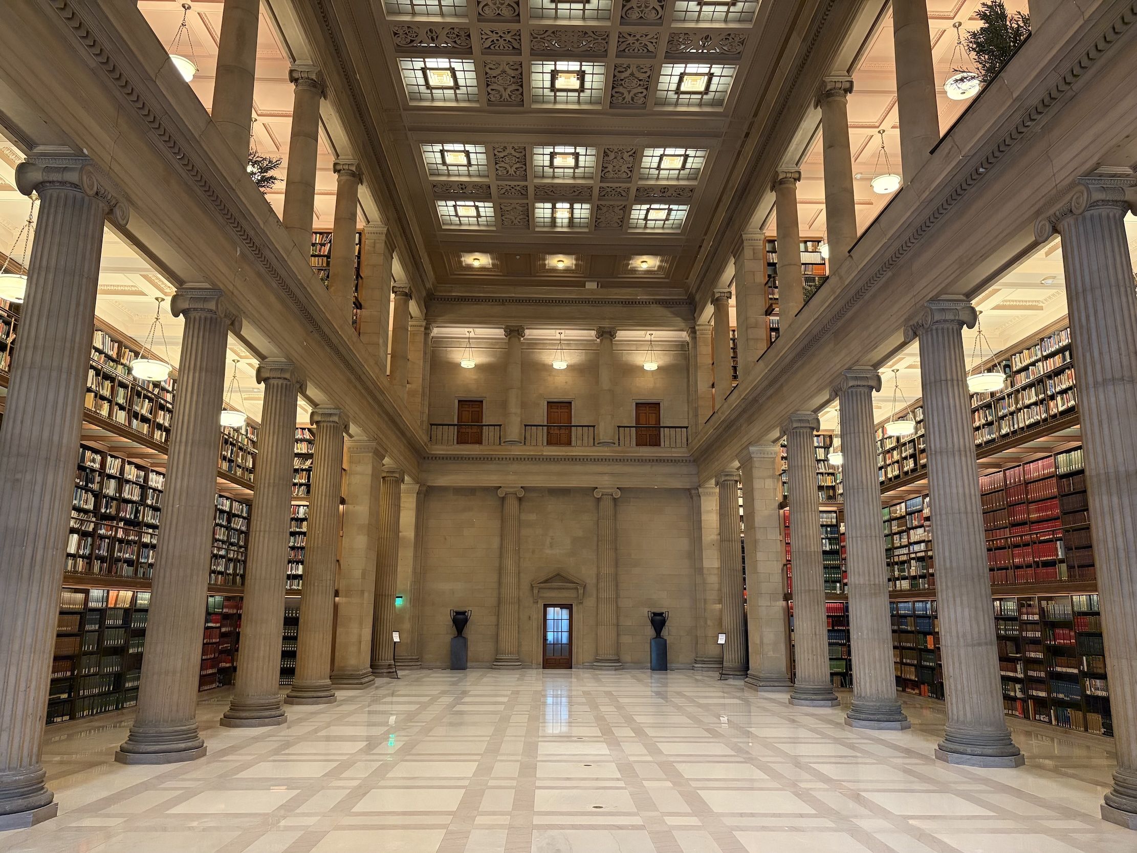 Grand neoclassical library interior with tall marble columns, two levels of bookshelves along the walls, balconies, and a checkered marble floor; warm lights illuminate the ornate ceiling.