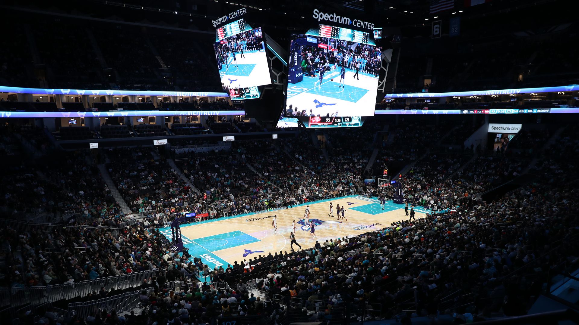 Wide shot of a basketball game in a crowded arena. A bright blue court with players mid-game; a large hanging scoreboard shows action and Spectrum Center branding, surrounded by fans.