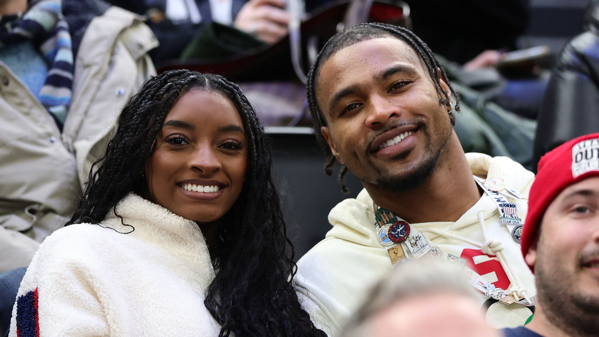 Two people sit in stands smiling at the camera: left, a woman with long braids in a cream fleece jacket; right, a man with cornrows in a light hoodie wearing pins, with blurred spectators behind.