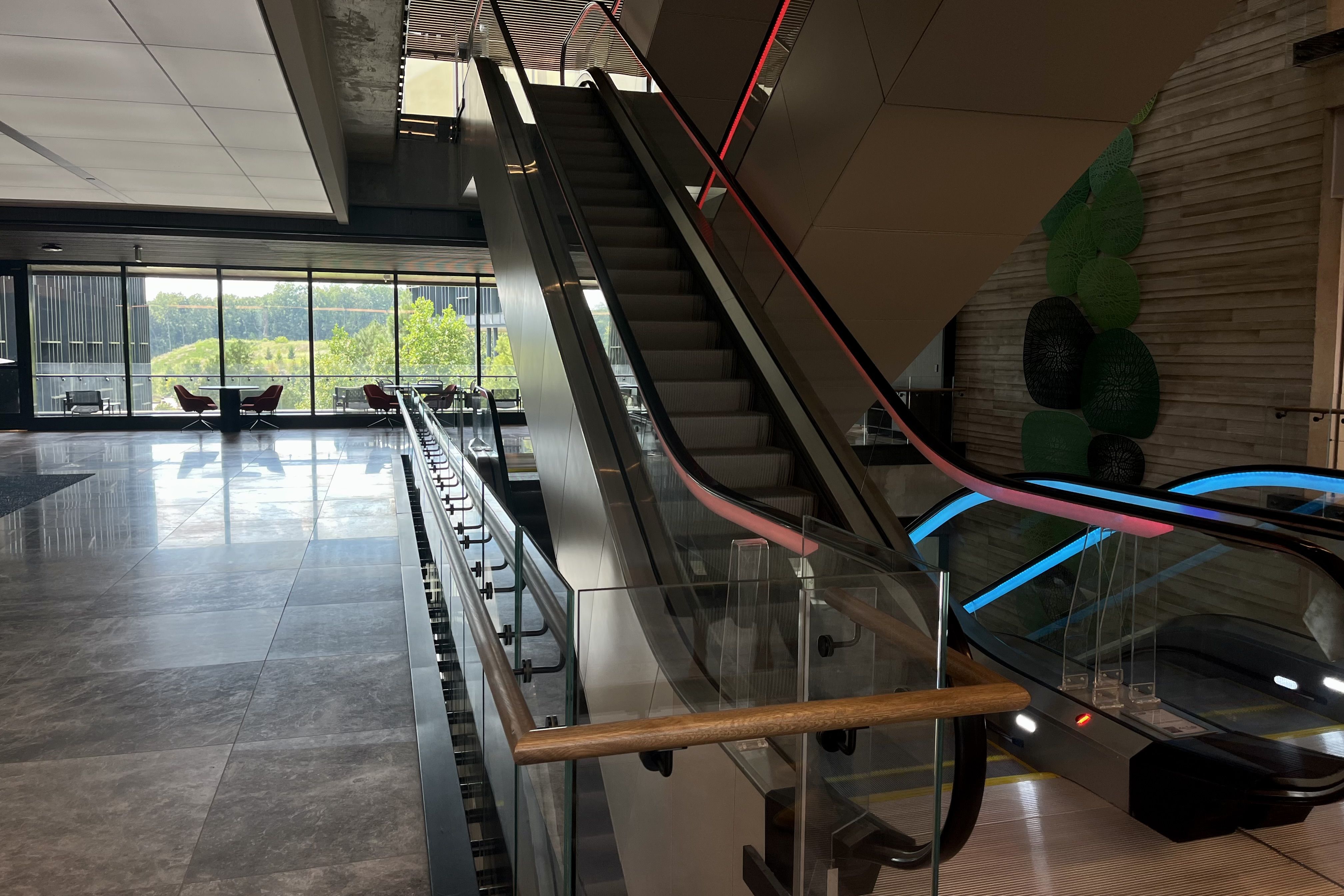 Indoor modern lobby with an empty escalator featuring red and blue handrail lights, glass railings, gray tile floor, and large windows showing greenery outside with chairs and tables.