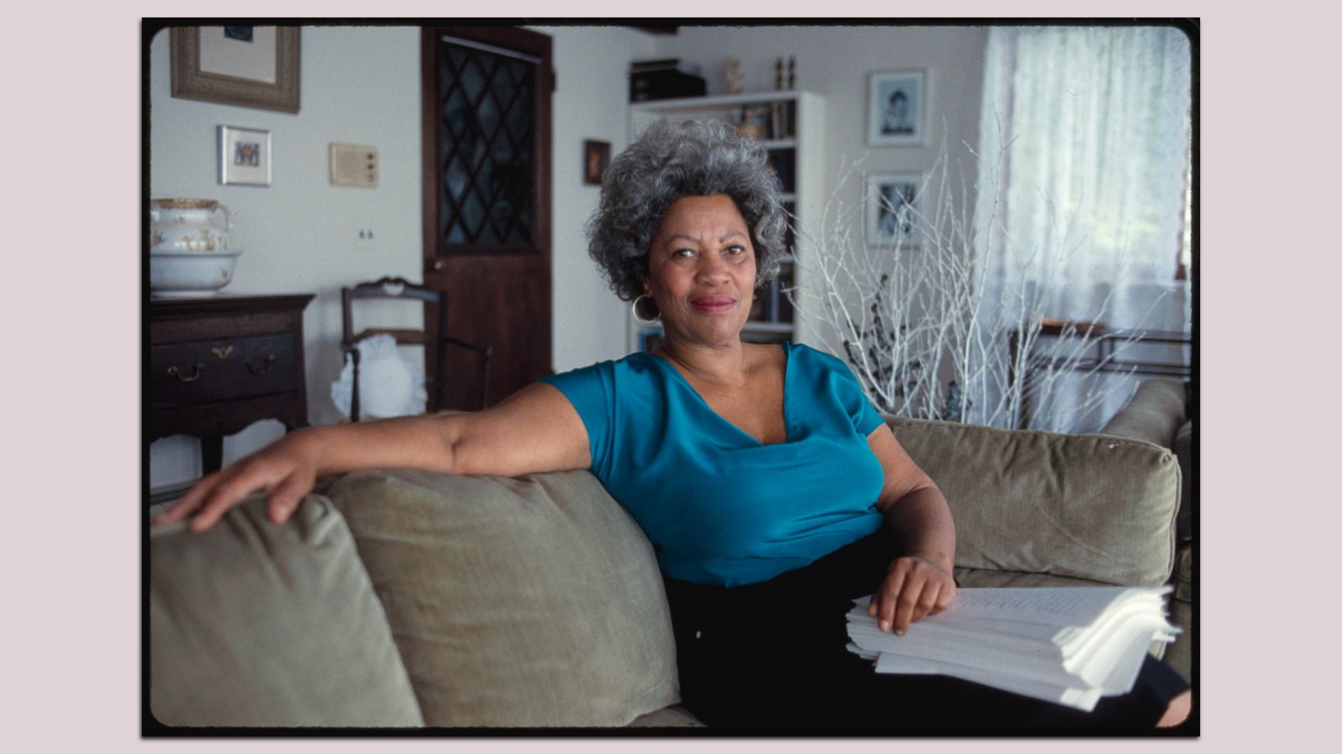 Woman with gray curly hair and blue top sitting on a beige couch in a cozy room, holding a stack of papers, with wooden furniture and framed pictures in the background.
