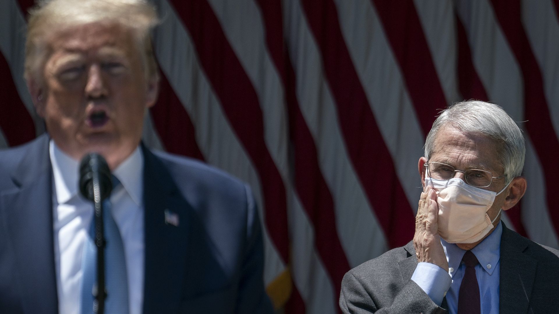 Photo of Dr. Anthony Fauci rubbing his masked face as he watches President Trump give a speech