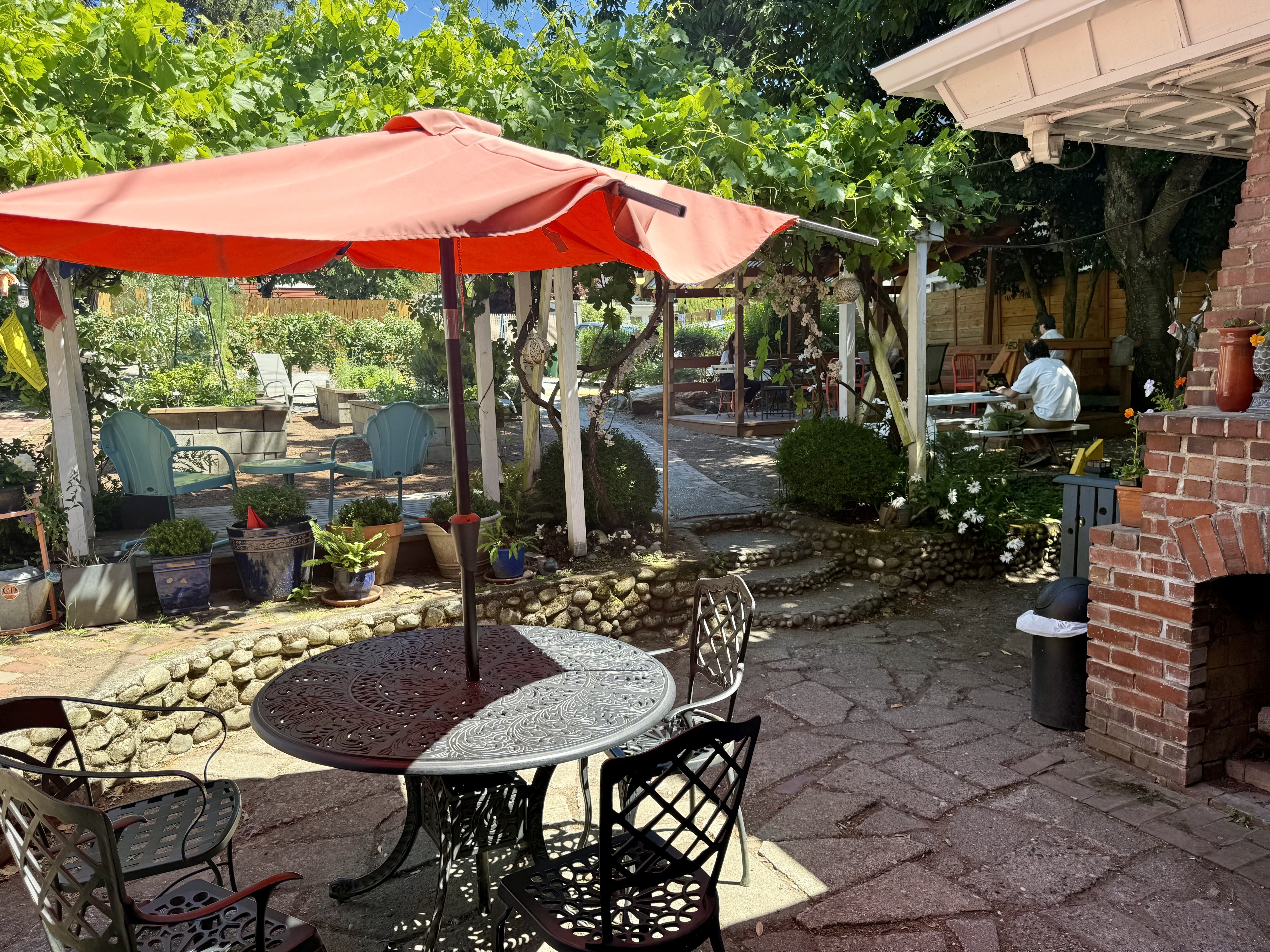 Sunny backyard patio with a red umbrella over metal tables and chairs, potted plants along stone borders, lush green trees, and people seated under a wooden pergola near a brick wall.