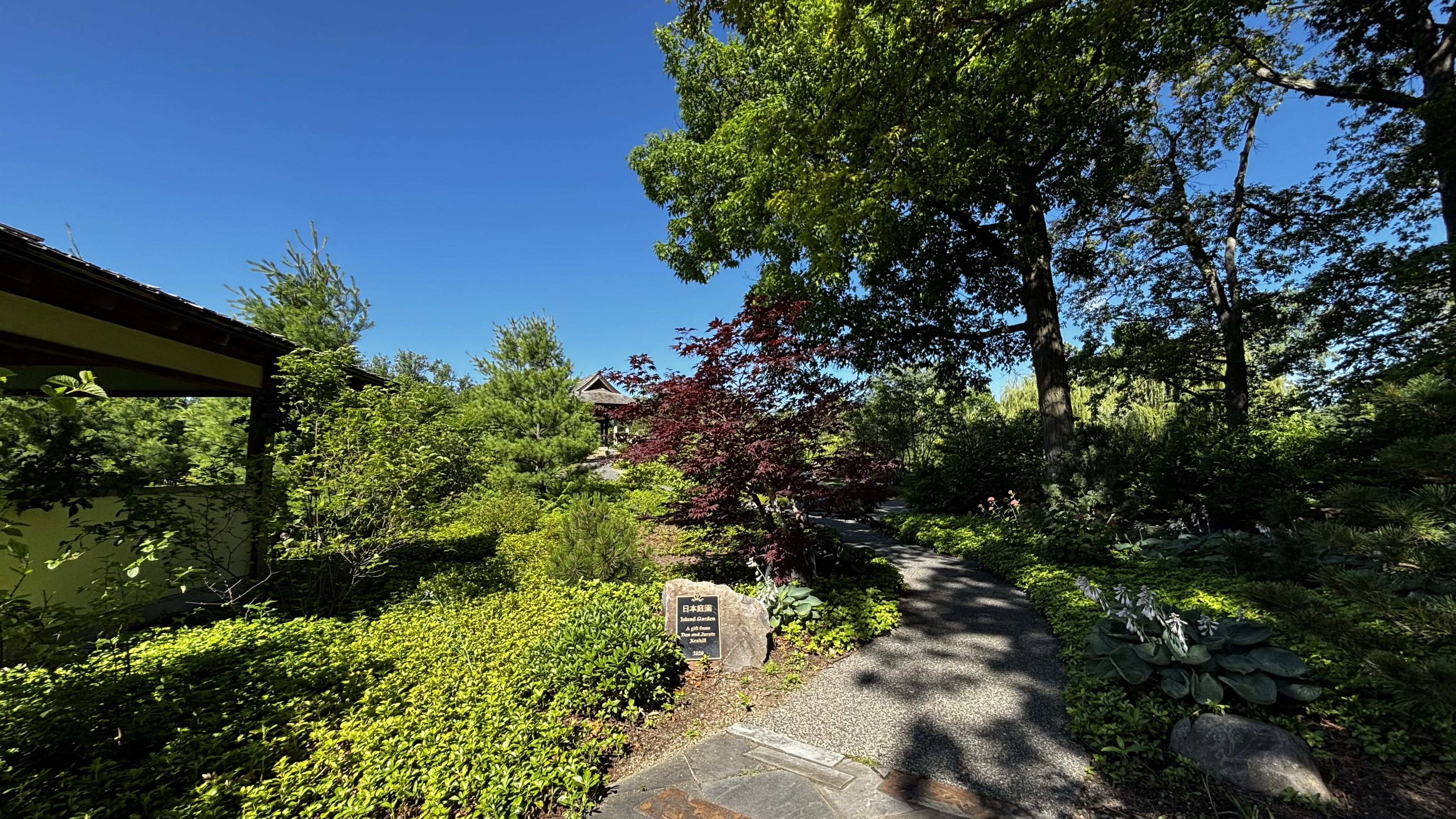 Sunny garden under a blue sky with lush trees and a gravel path. A yellow-walled building with a dark roof sits on the left, and a red-leaf tree adds color near a stone plaque by the path.