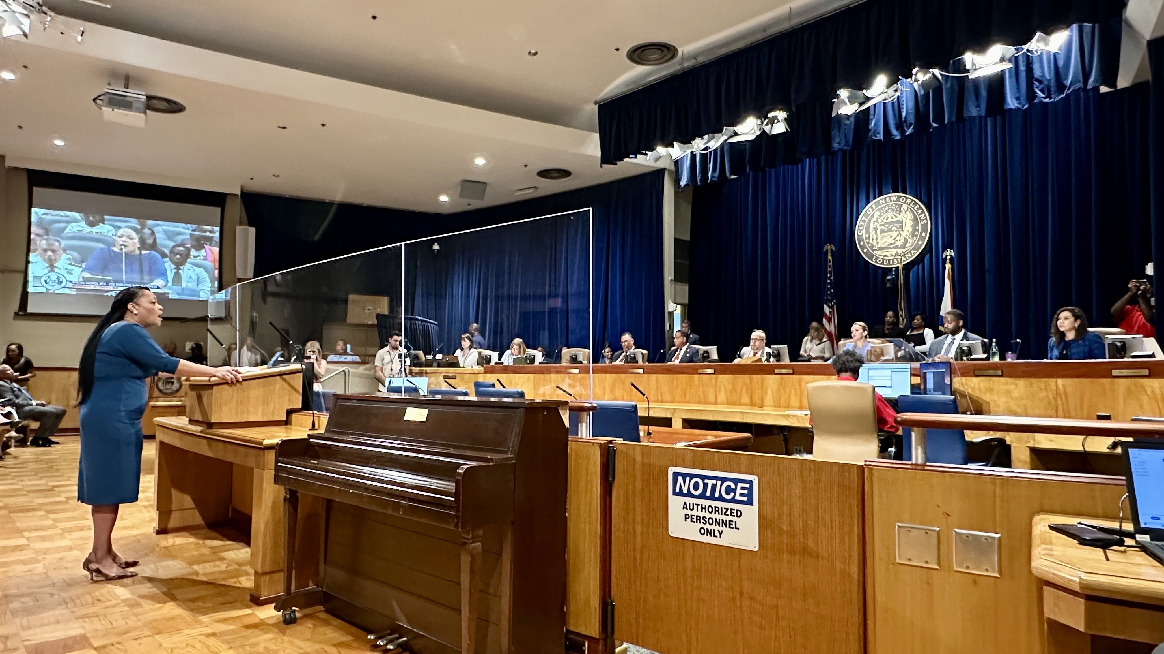 Woman in blue dress speaking at podium in a council chamber with officials seated in a semi-circle. City of New Orleans seal and flags are visible on dark blue curtains behind them.