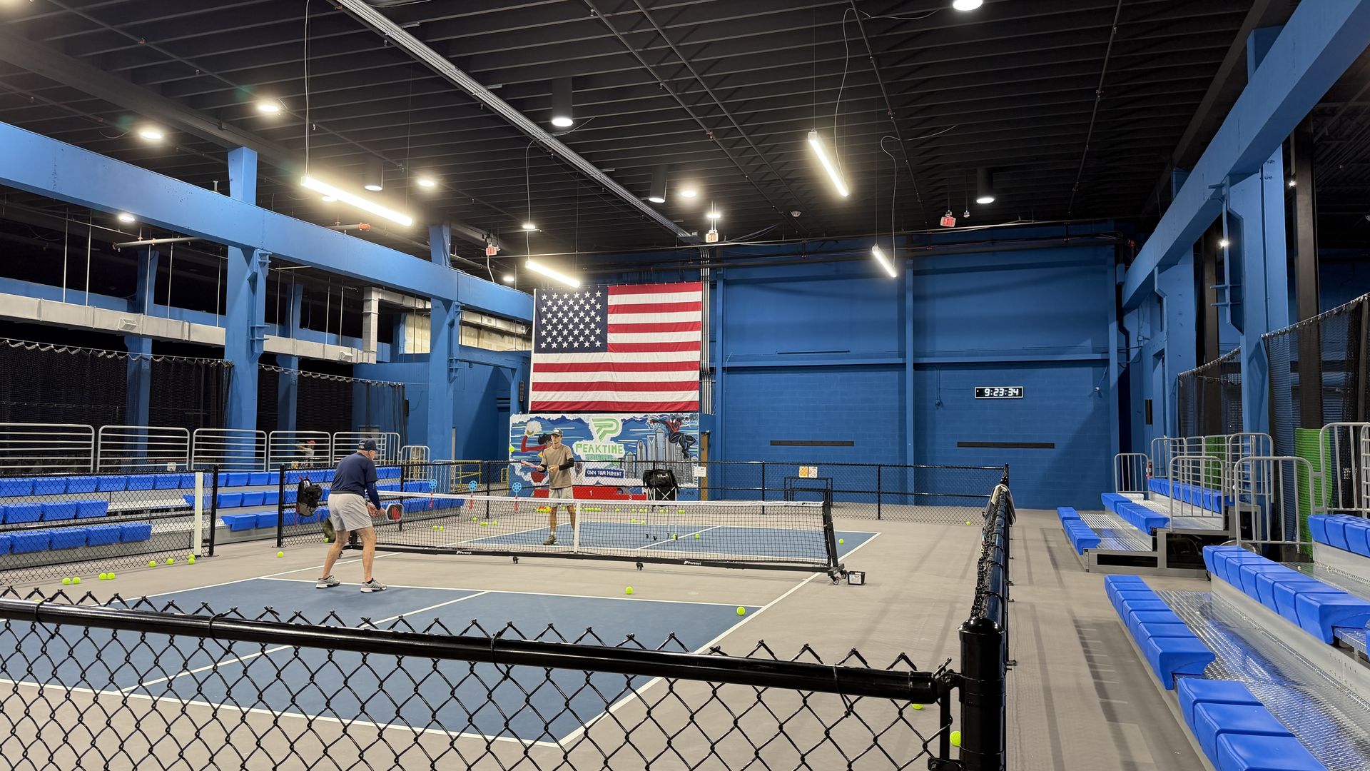 Indoor tennis court with blue walls and beams. A large American flag hangs behind the back wall; two players near the net, tennis balls scattered, blue bleachers on either side.