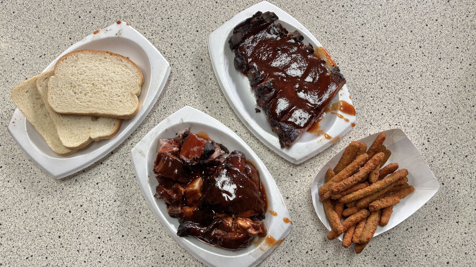 Overhead shot of a BBQ meal on foam trays: sliced white bread on the left; glossy, saucy ribs on the right; chopped rib pieces in sauce on a bottom tray; and a plate of seasoned fried sticks.