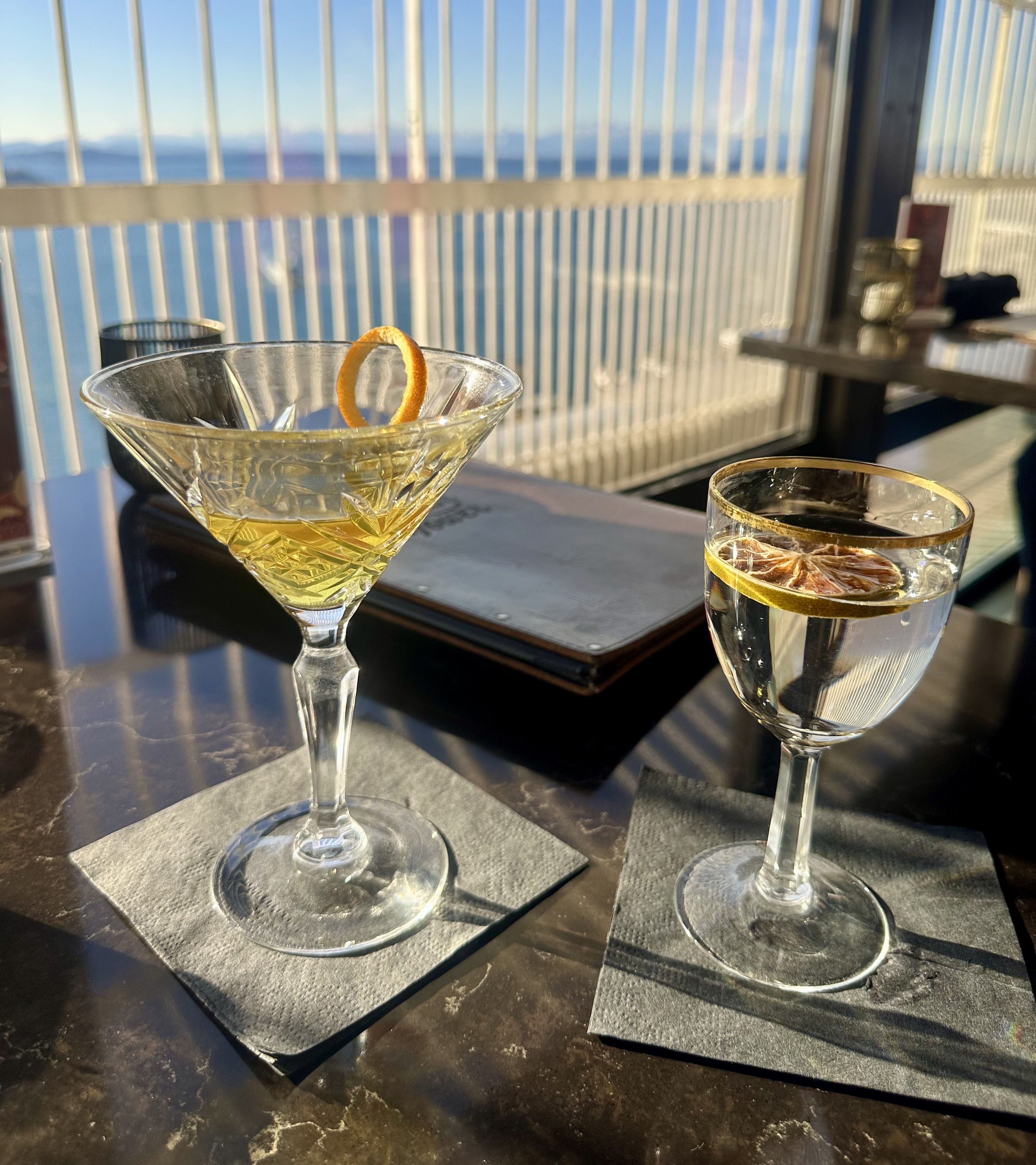 Two drinks sitting on a table in front of a large window with a view of sky and ocean. One drink is in a martini-style glass, the other in a small goblet, both with citrus garnishments.